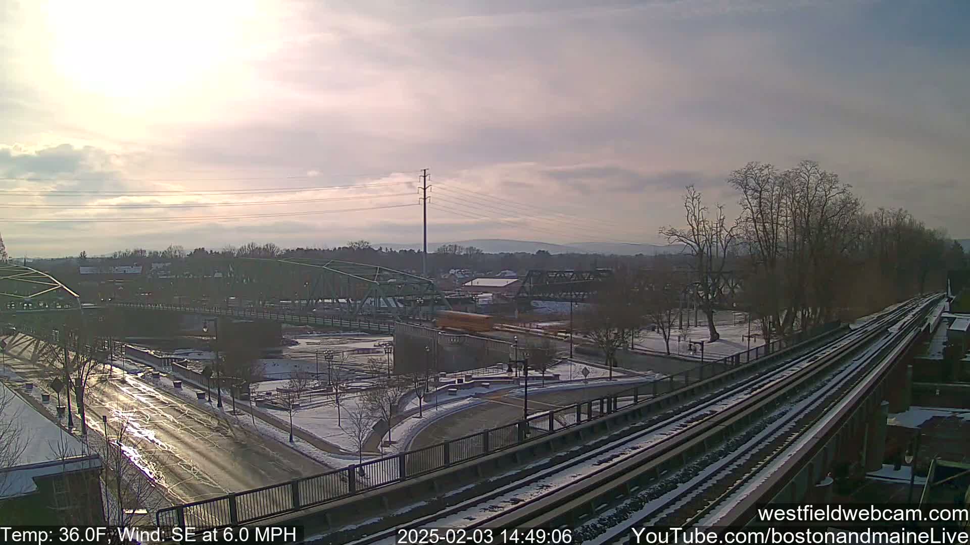 A partly sunny, 36-degree day with a southeast wind at 6.0 mph reveals a snow-dusted landscape featuring two bridges, train tracks, and a road, with trees lining the riverbank in the background.