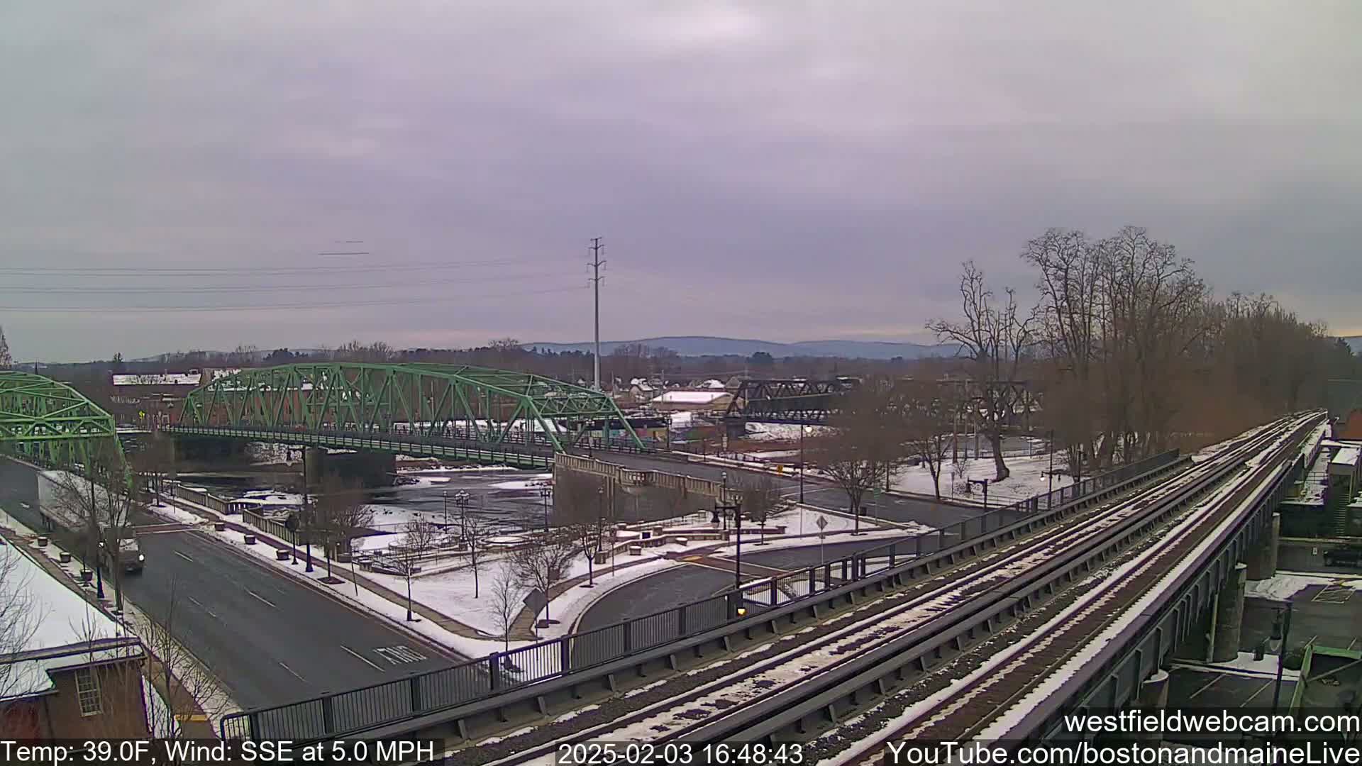 A green bridge spans a river, with train tracks and a snow-covered roadway visible in the foreground, under an overcast sky.