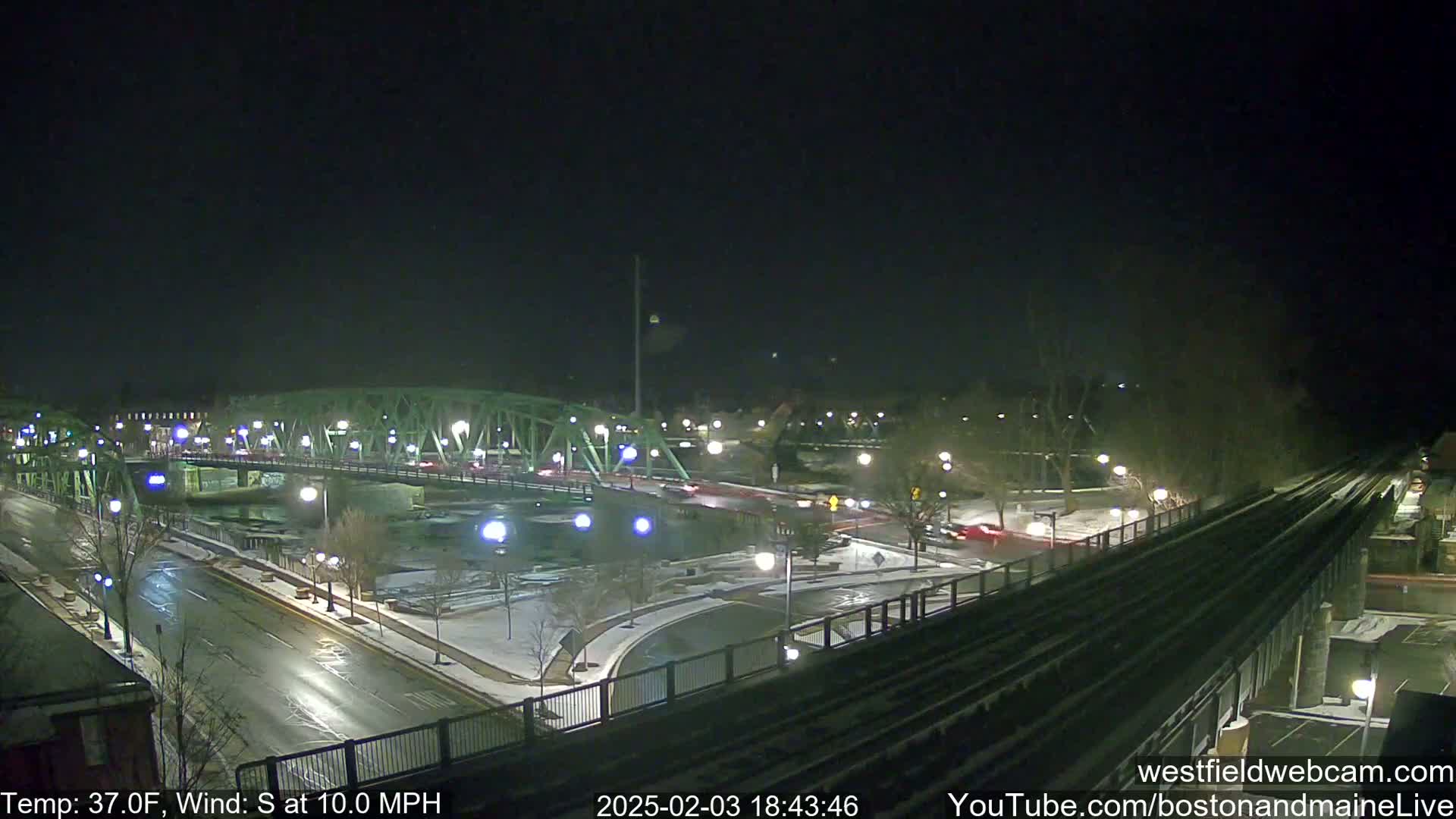 A nighttime view of a bridge and roadway intersection with light snow on the ground, under a clear dark sky, with a south wind at 10 mph.