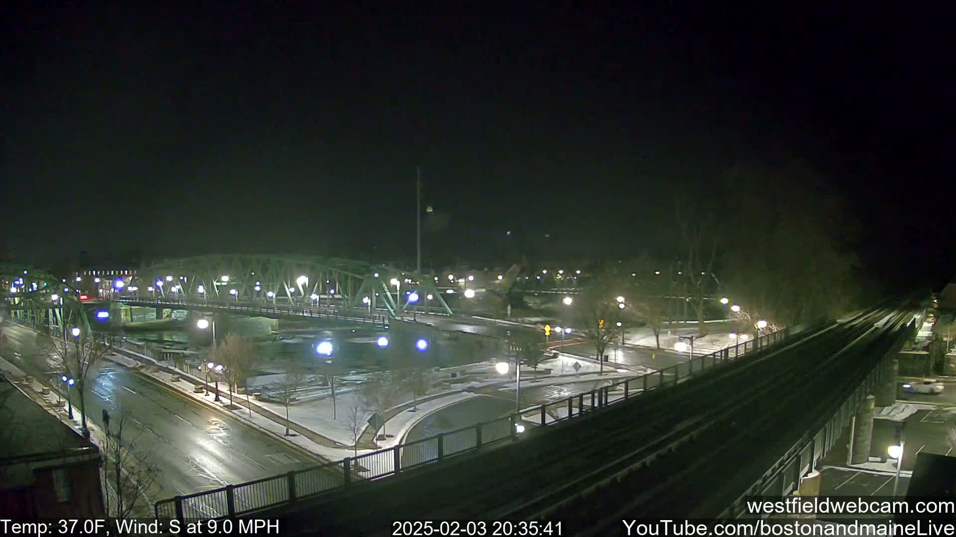 A nighttime view of a bridge over a river, with a road and train tracks, under partly snowy conditions and a south wind.