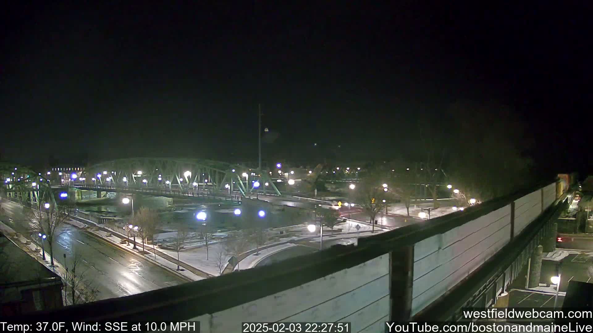 A nighttime view of a bridge over a river, with a road and some snow on the ground, under a dark sky with a 37°F temperature and a 10 mph wind from the south-southeast.