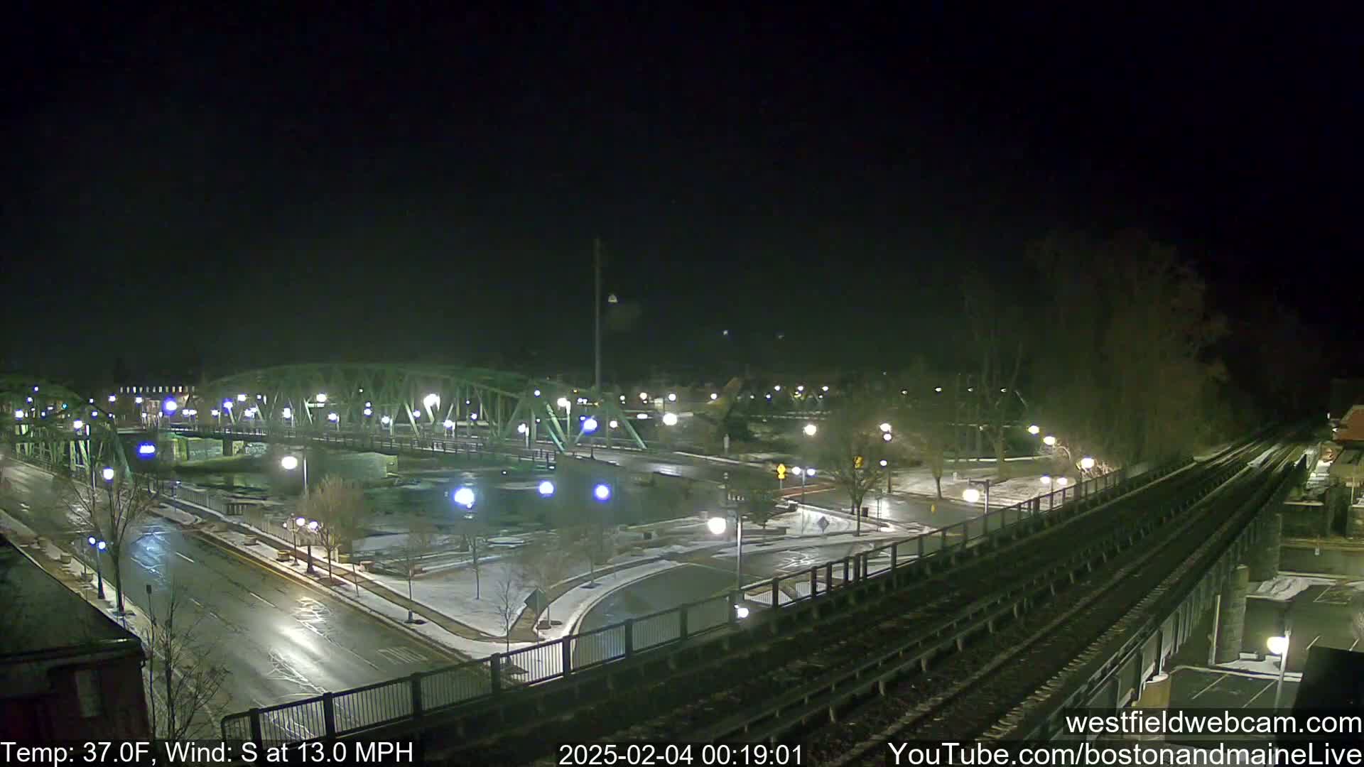 A nighttime view of a bridge and roadway, partially covered in snow, with a railway in the foreground under a dark sky and a south wind at 13 mph.