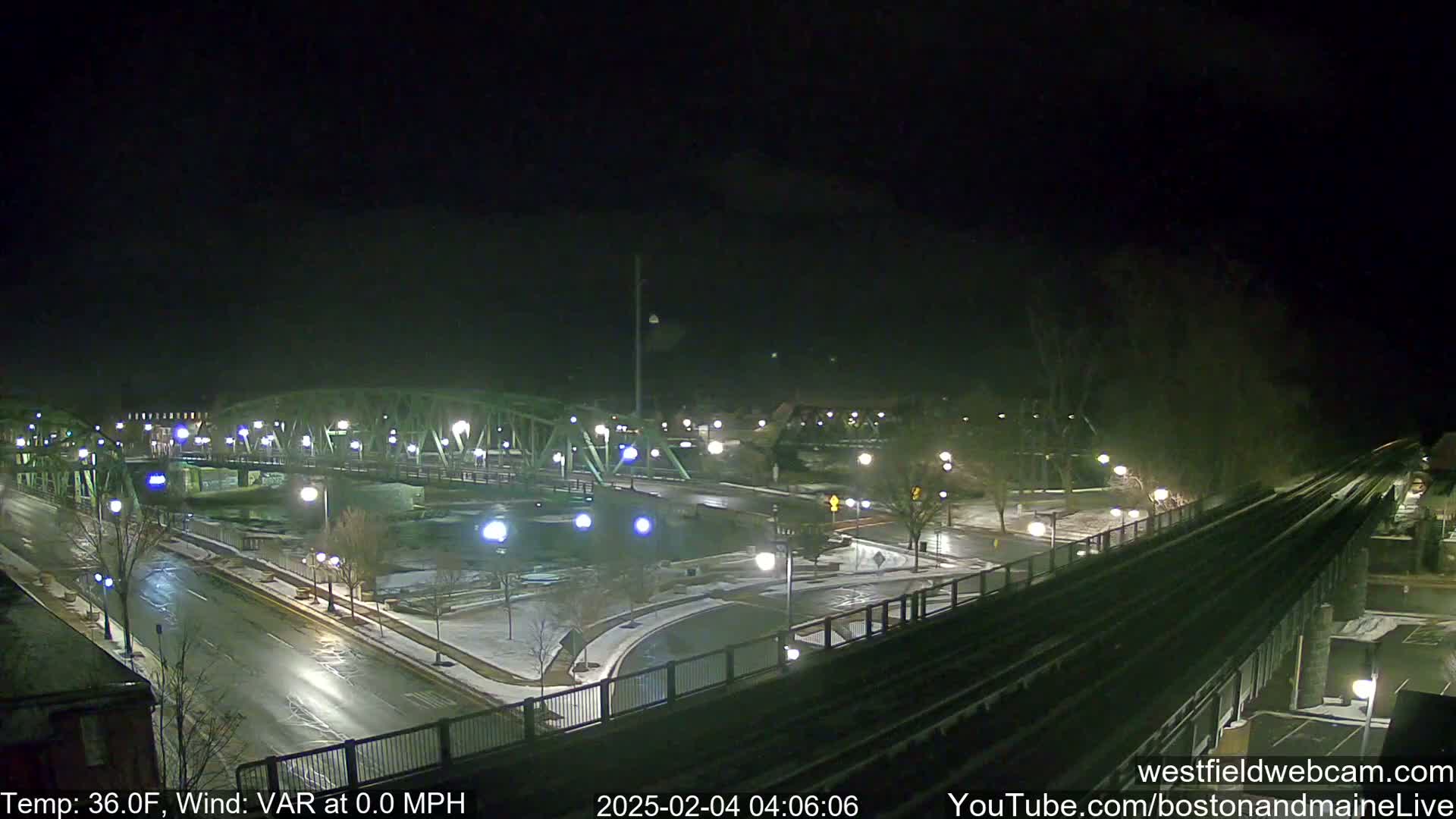 A nighttime view of a bridge over a river, with snow on the ground and wet roads, under a dark sky.