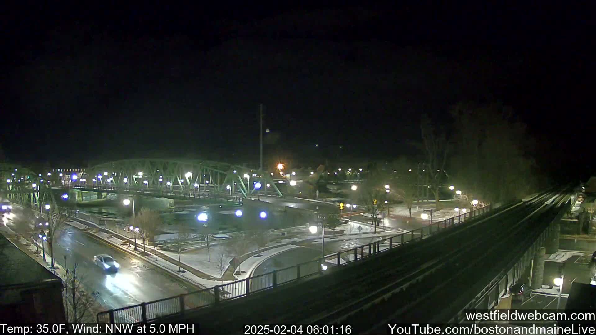 A nighttime view of a bridge and roadway with light snow on the ground, under a dark sky with a north-northwest wind at 5 mph and a temperature of 35 degrees Fahrenheit.