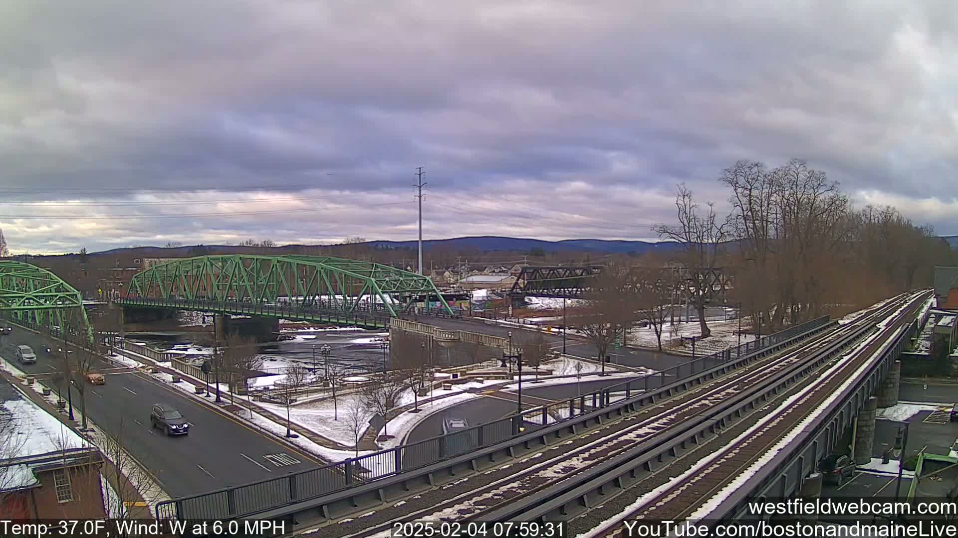A partly cloudy, cool day reveals a view of two bridges over a river, with a train track running alongside and some light snow on the ground.