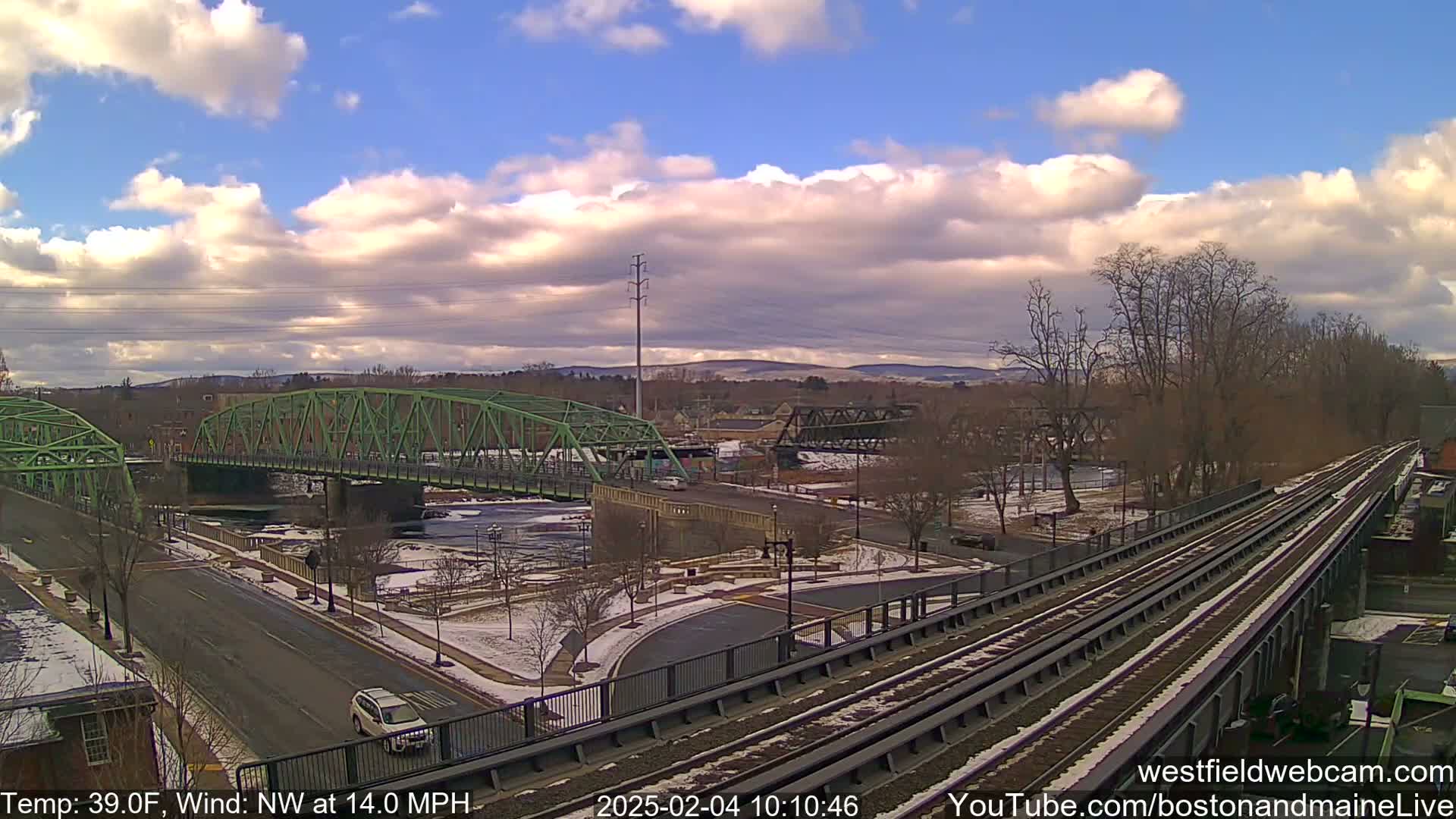 A partly cloudy sky over a town with two bridges, one green, the other dark metal, and train tracks running through a snowy landscape.