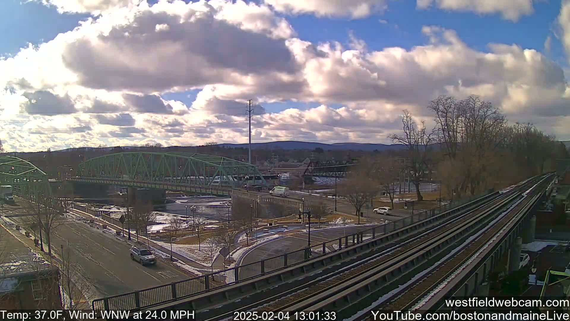 A partly cloudy day with a strong wind shows a view of a river, bridge, and train tracks with light traffic.