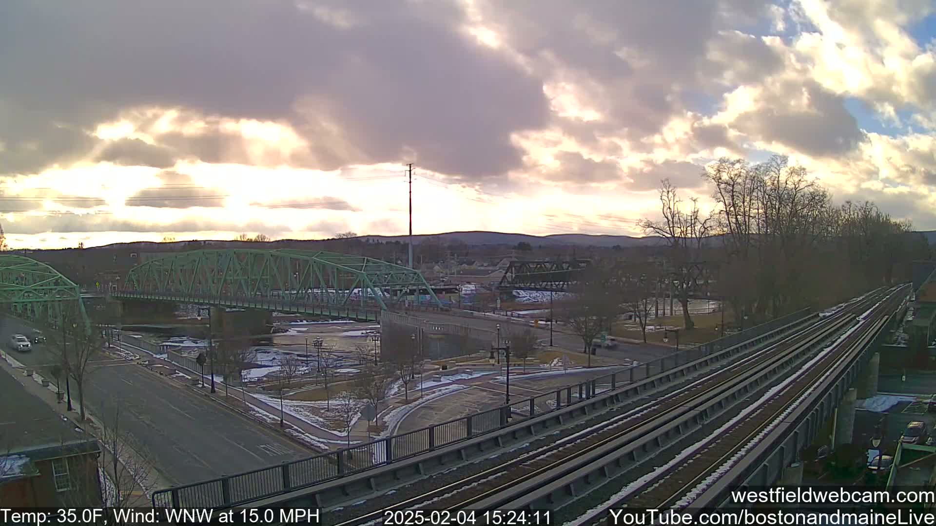 A partly cloudy sunset illuminates a cityscape featuring a green bridge over a river, a train track, and patches of snow on the ground.