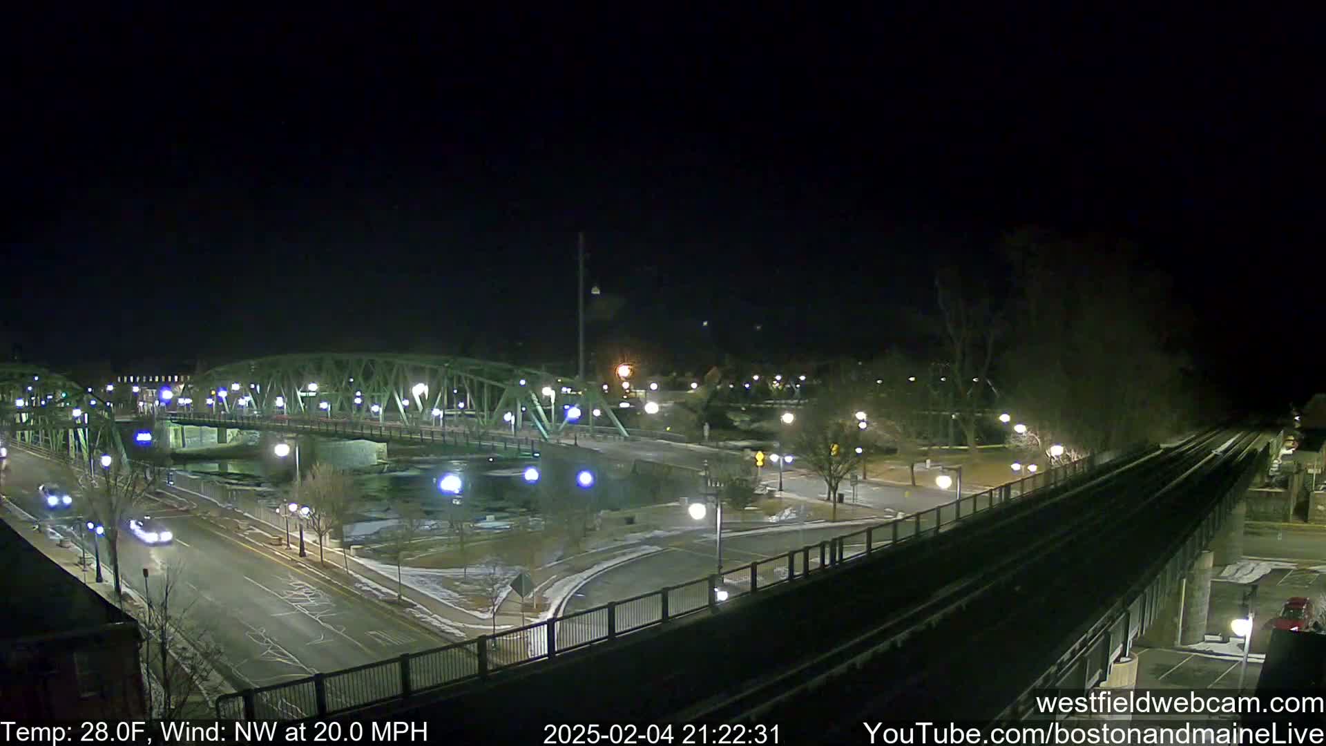 A nighttime, illuminated view of a bridge over a river, with a roadway and train tracks visible, under a dark sky with a northwest wind of 20 mph and a temperature of 28°F.