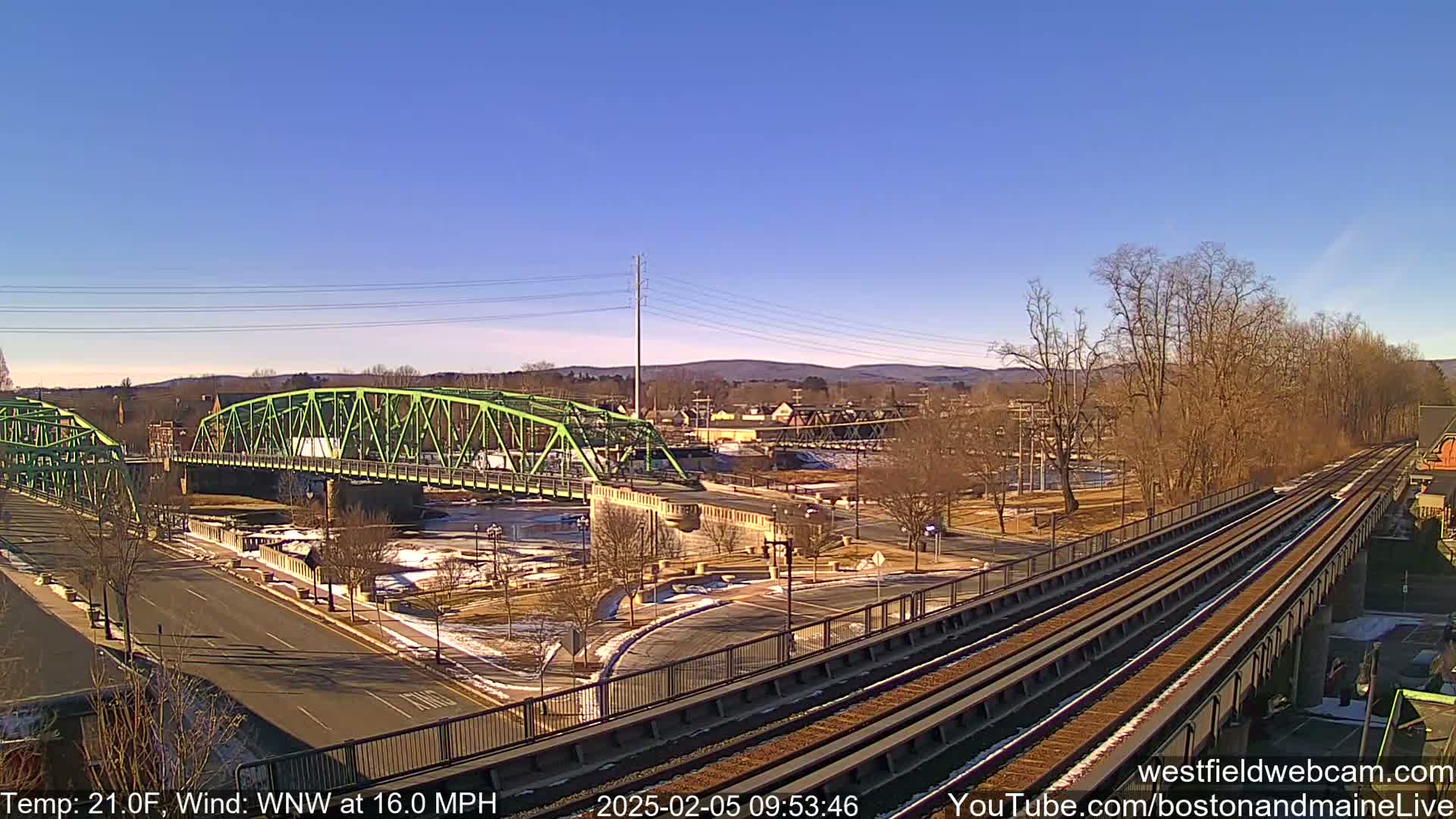 A green bridge spans a river, with a train track and roadway intersecting nearby under a clear, sunny sky.