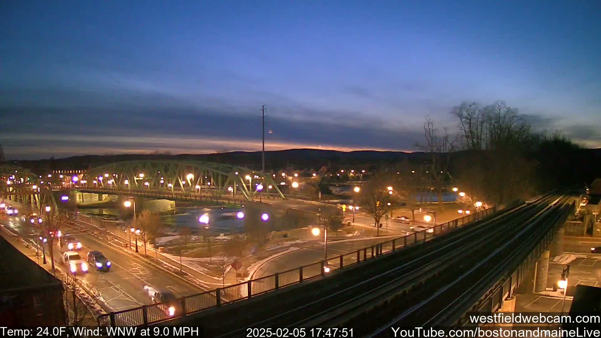 A nighttime view of a bridge over a river, with cars on the road below and train tracks in the foreground, under a mostly clear twilight sky, with a temperature of 24°F and a wind blowing from the west-northwest at 9.0 mph.