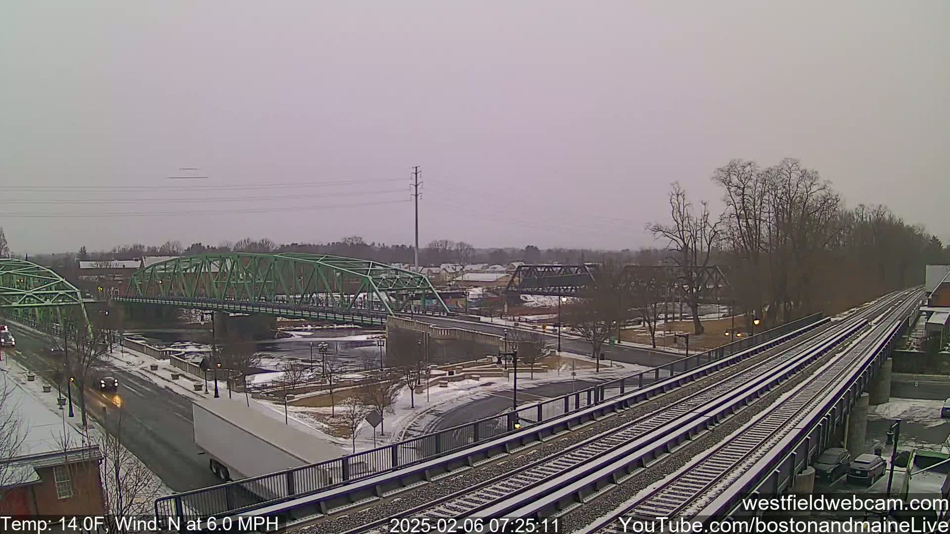 A view of a snow-covered town with two bridges over a river, train tracks, and a partially visible roadway.
