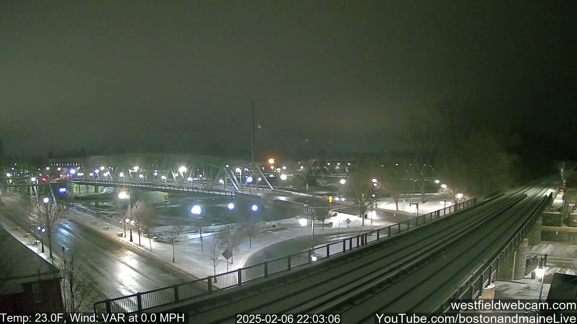 A snow-covered bridge and roadway are illuminated at night by streetlights, with train tracks visible in the foreground.