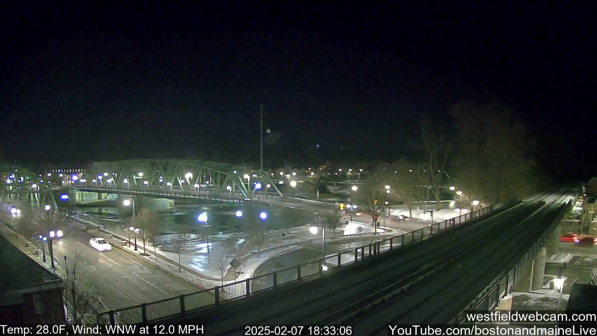 A nighttime view of a bridge over a river, with a road and train tracks, all lightly covered in snow, under a clear sky with a wind of 12 mph.