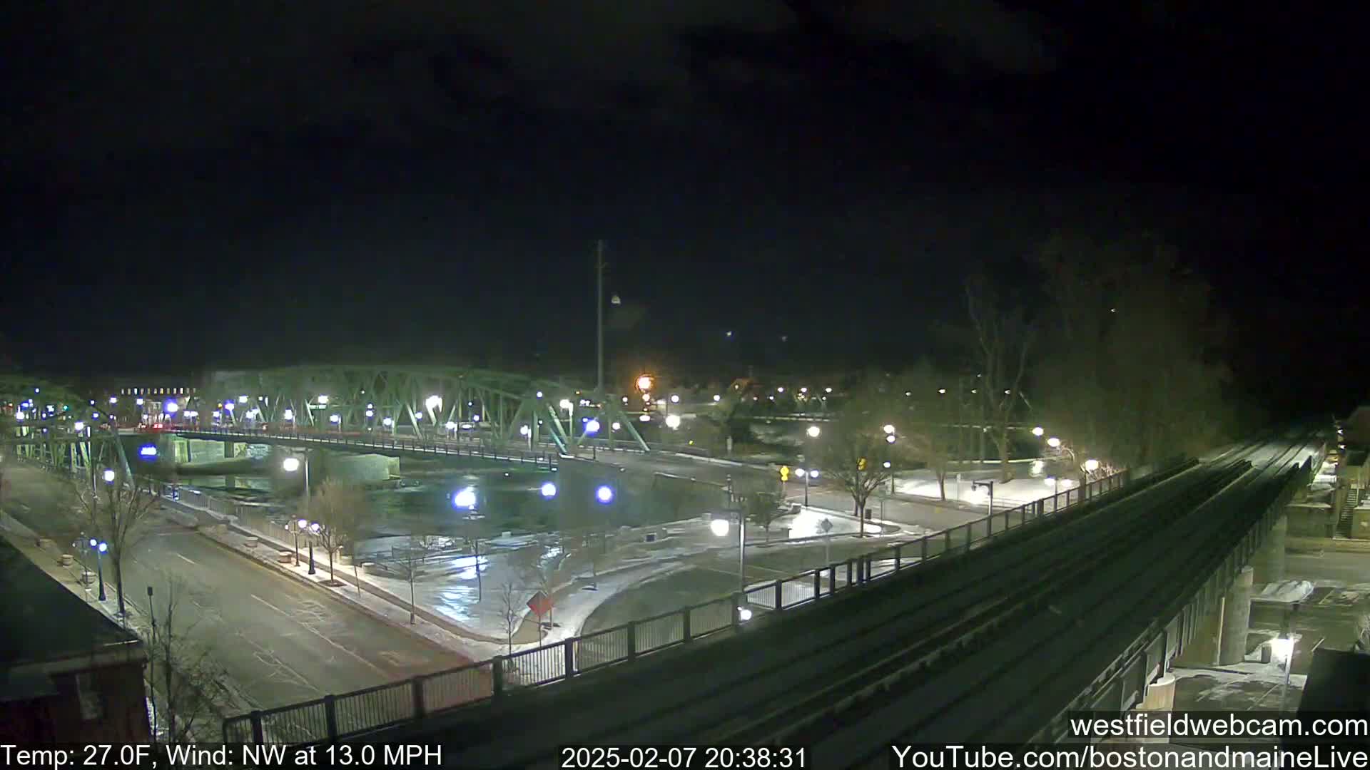 A nighttime view of a bridge over a river, with a train track alongside a snow-dusted road, under a dark, windy sky with a temperature of 27 degrees Fahrenheit.