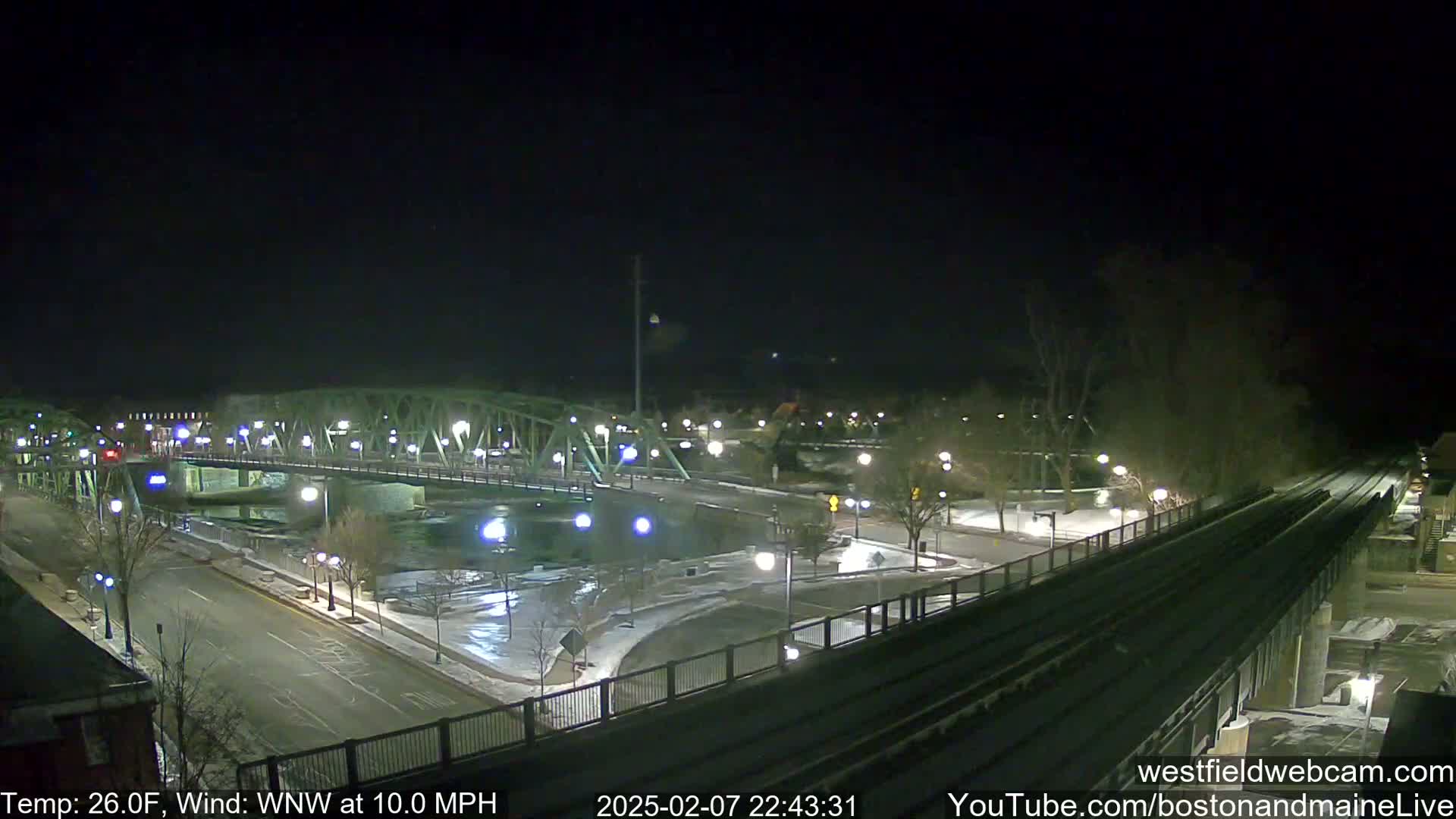 A nighttime view of a bridge over a river, with a train track and roadway in the foreground, under a clear sky with a temperature of 26°F and a wind of 10 mph from the west-northwest.