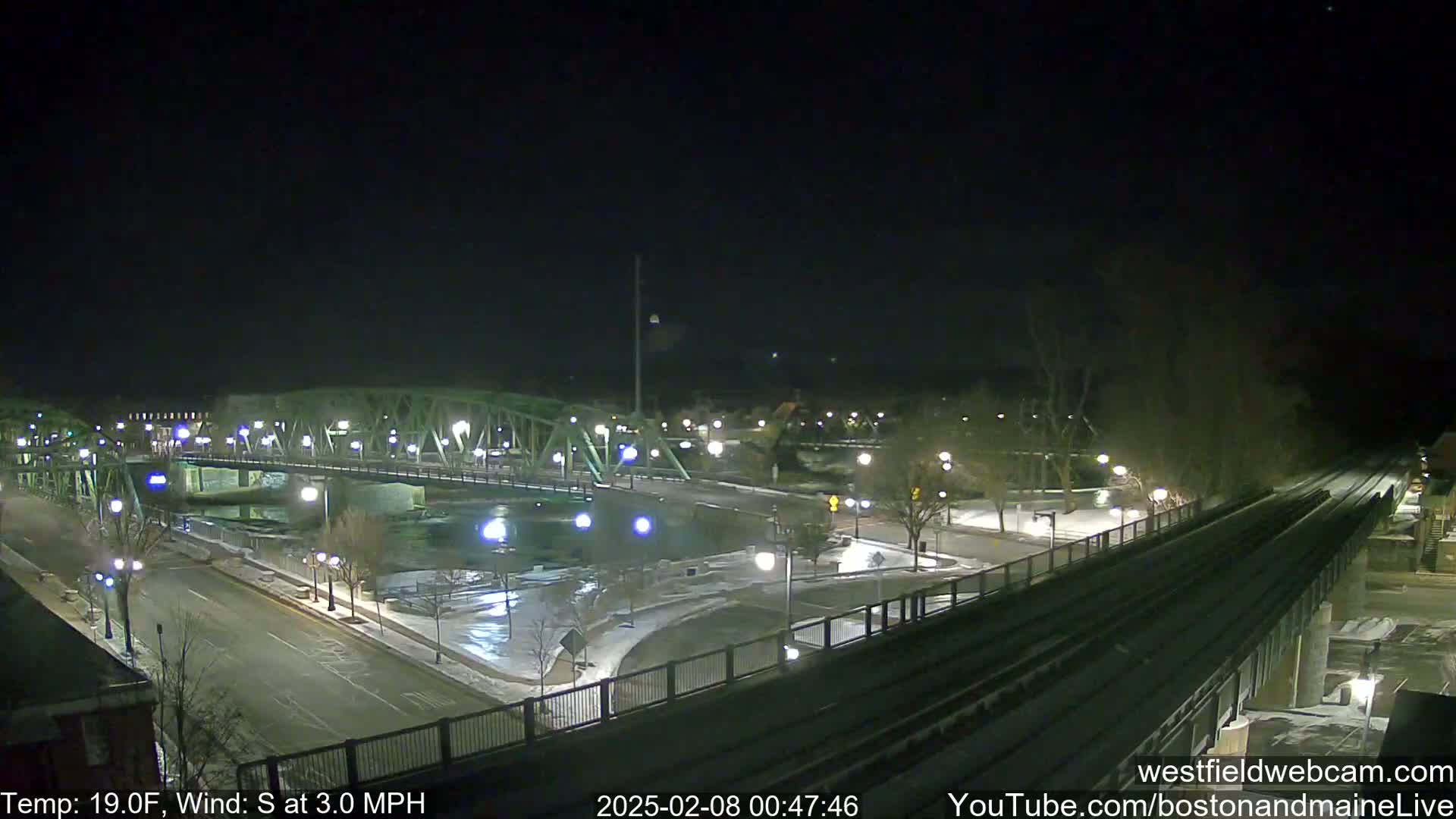 A nighttime view of a bridge over a river, with snow on the ground and a light dusting of snow on train tracks, under a clear, dark sky.