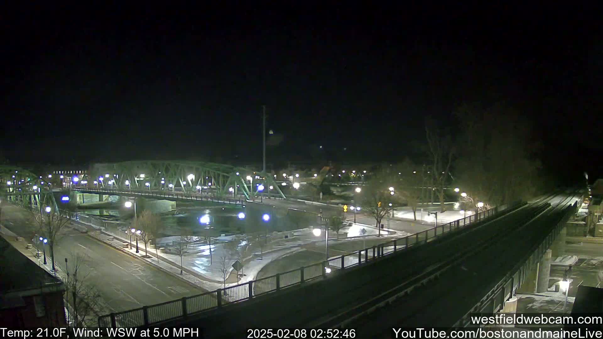 A nighttime view of a bridge over a river, with snow on the ground and a train track visible in the foreground, under a clear, dark sky.