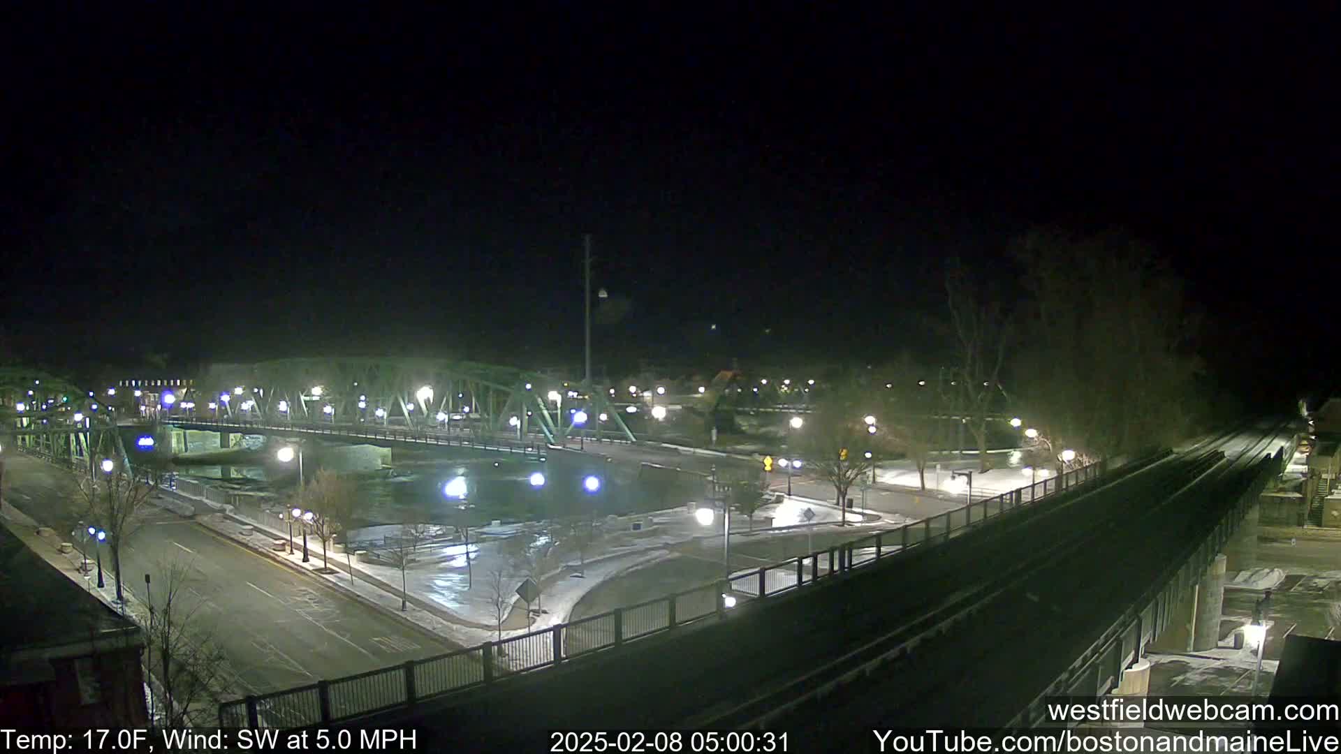 A nighttime view of a bridge over a river, with some snow on the ground and a southwest wind.