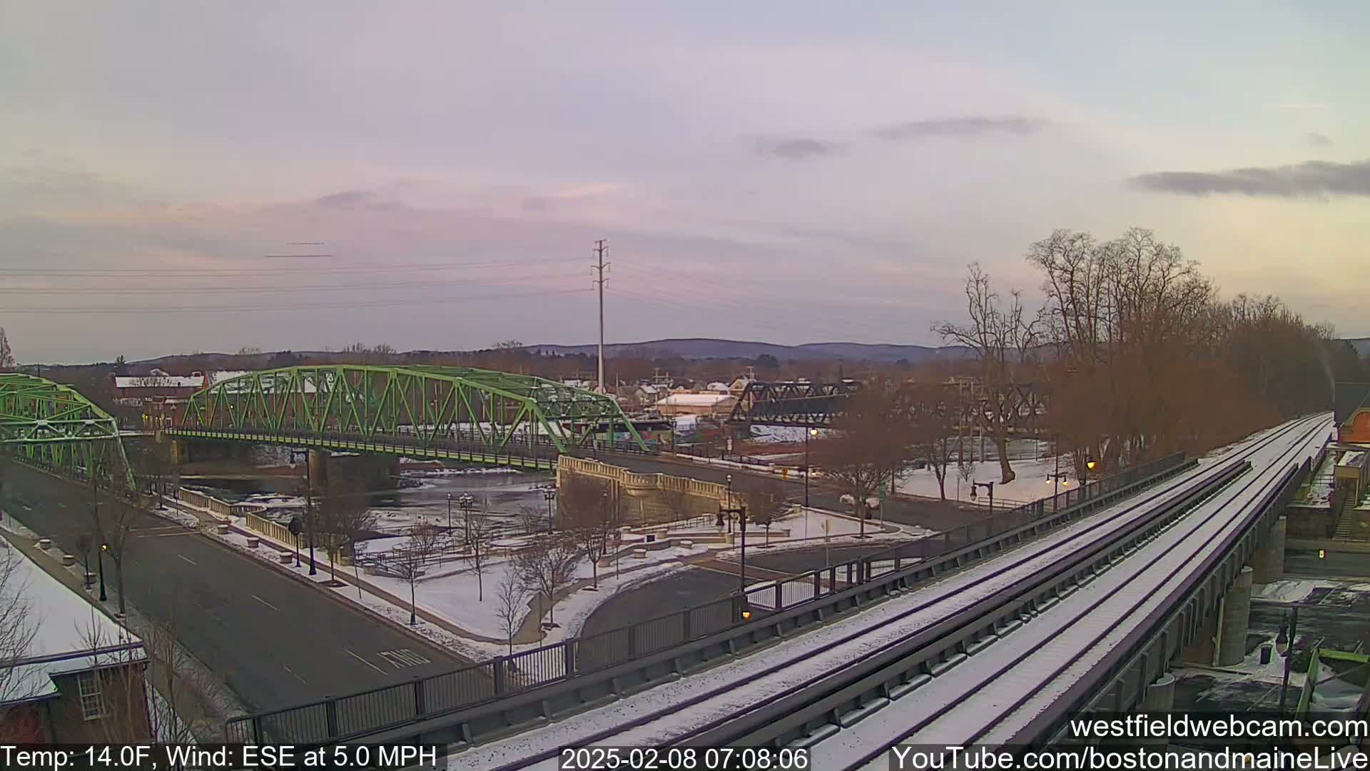 A snow-covered train track and a green bridge span a river in a town under a pale pink sky.