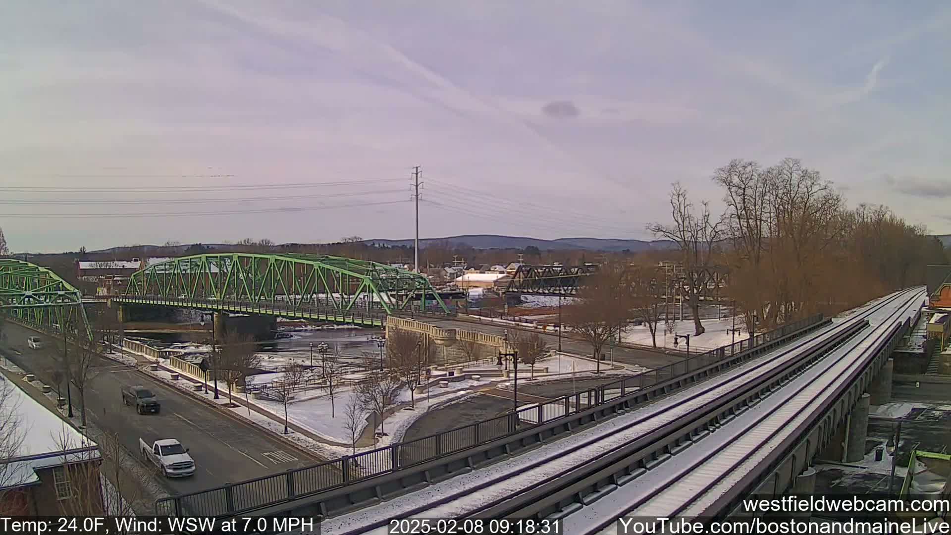 A light dusting of snow covers a town scene with two bridges spanning a river, one green and one dark metal, viewed from an elevated perspective of a train track.