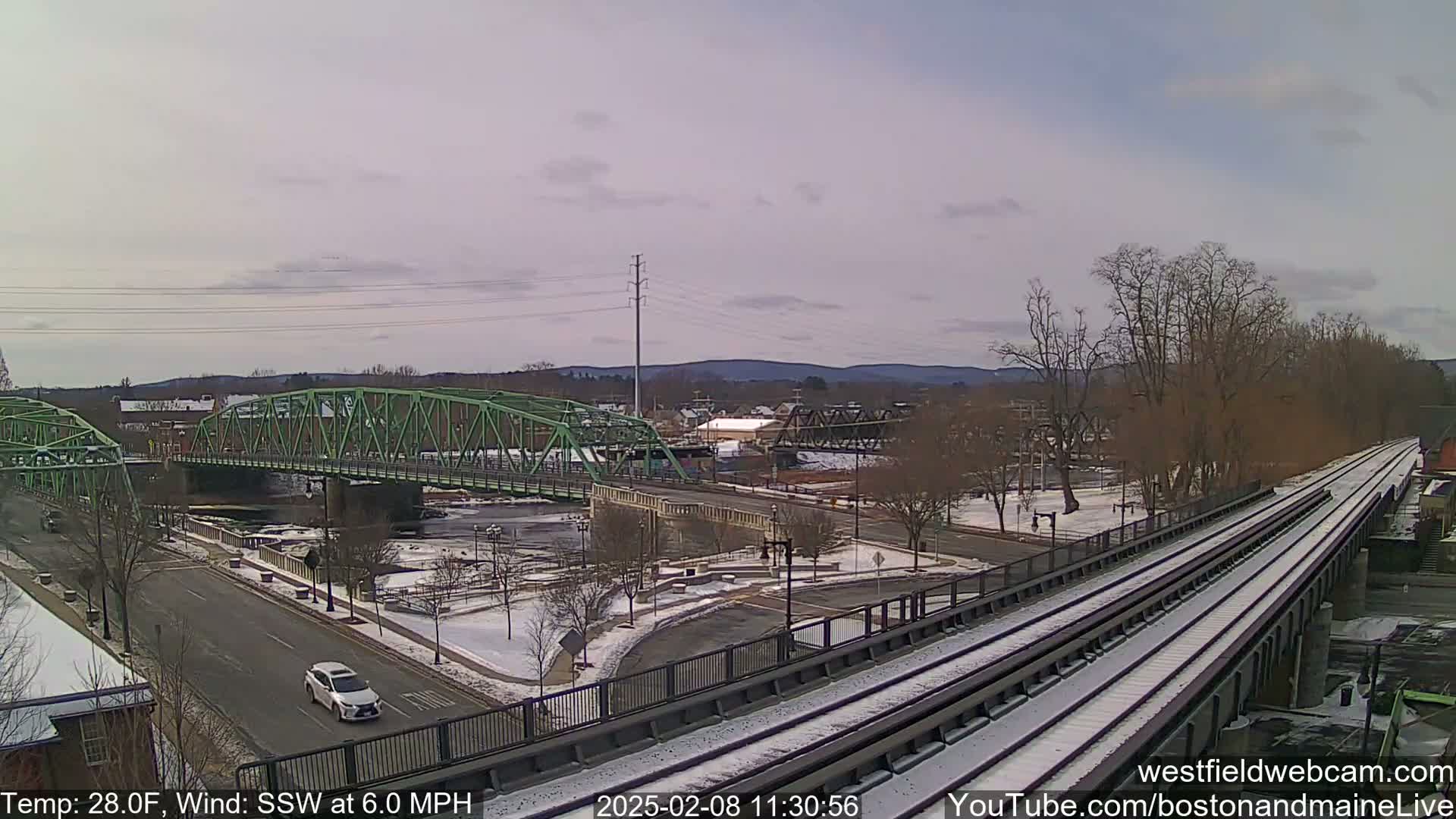 A light-green bridge spans a river in a snow-dusted town under a cloudy sky.
