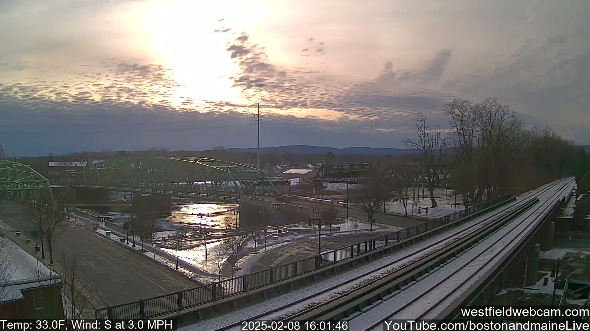 A partially snowy scene at sunset shows a river with two bridges, one green and one dark metal, and a train track in the foreground.