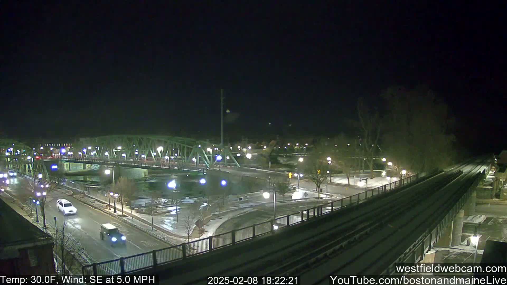 A nighttime view of a bridge over a river, with a train track and roads visible, and light snow cover on the ground under a dark sky, with a temperature of 30°F and a southeasterly wind of 5 mph.