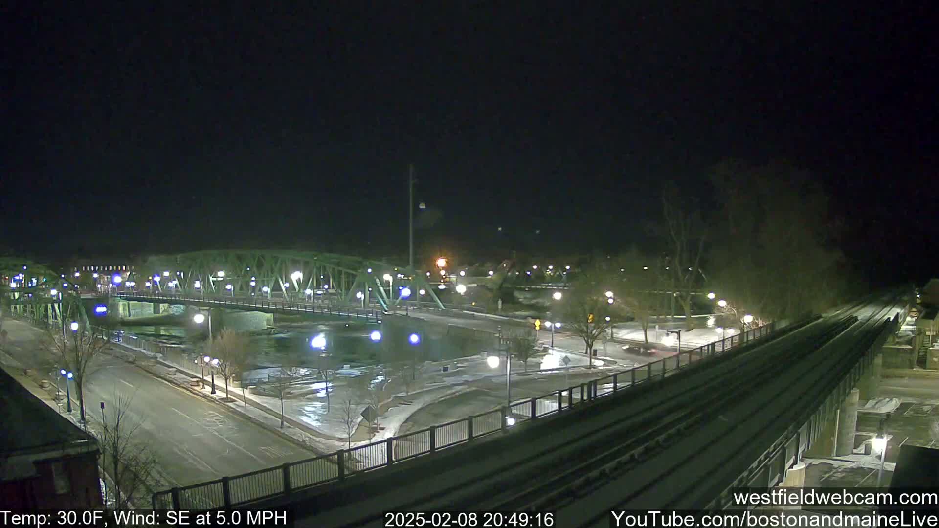 A nighttime view of a snow-covered bridge and river, with a train track and road visible, under a clear, dark sky.