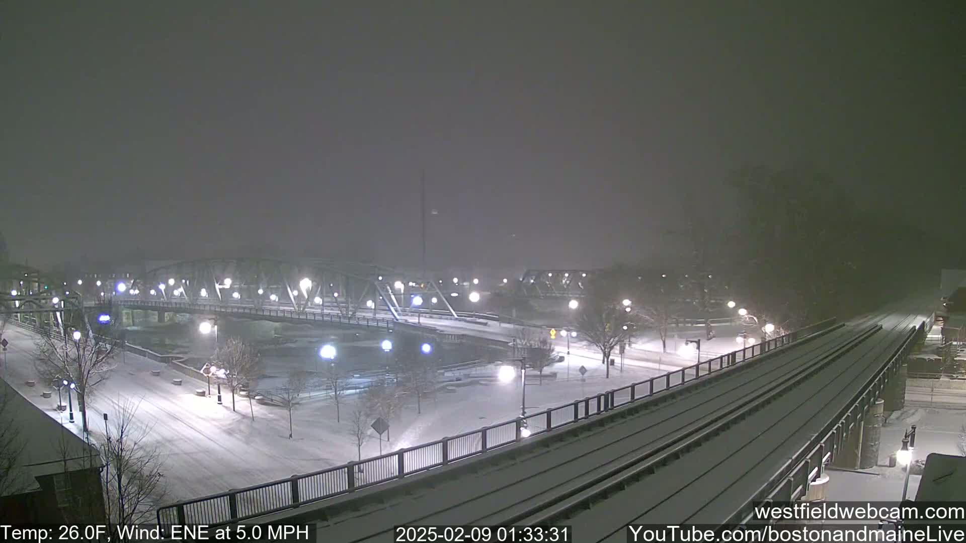 A snow-covered cityscape at night, featuring a bridge over a river and train tracks, with light snowfall and a wind of 5.0 mph.
