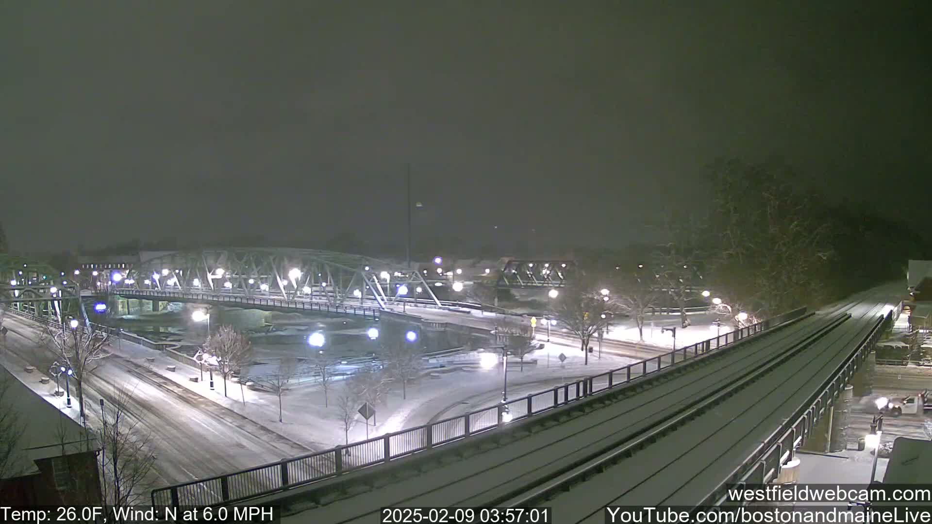 A snow-covered bridge and roadway are visible at night, with a train track running alongside, under a lightly falling snow.