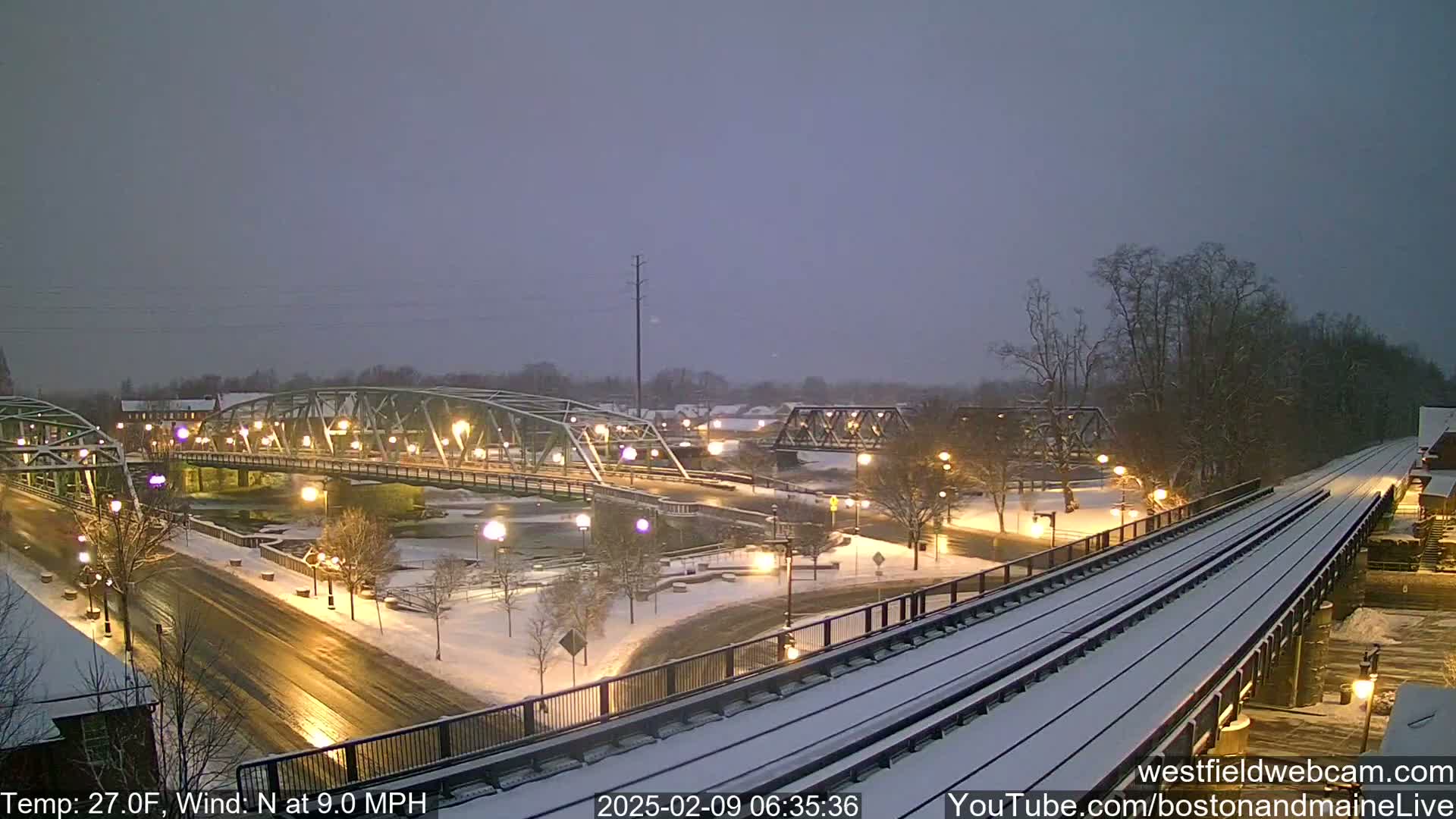 A snow-covered cityscape at night features two bridges over a river, illuminated streets, and train tracks.