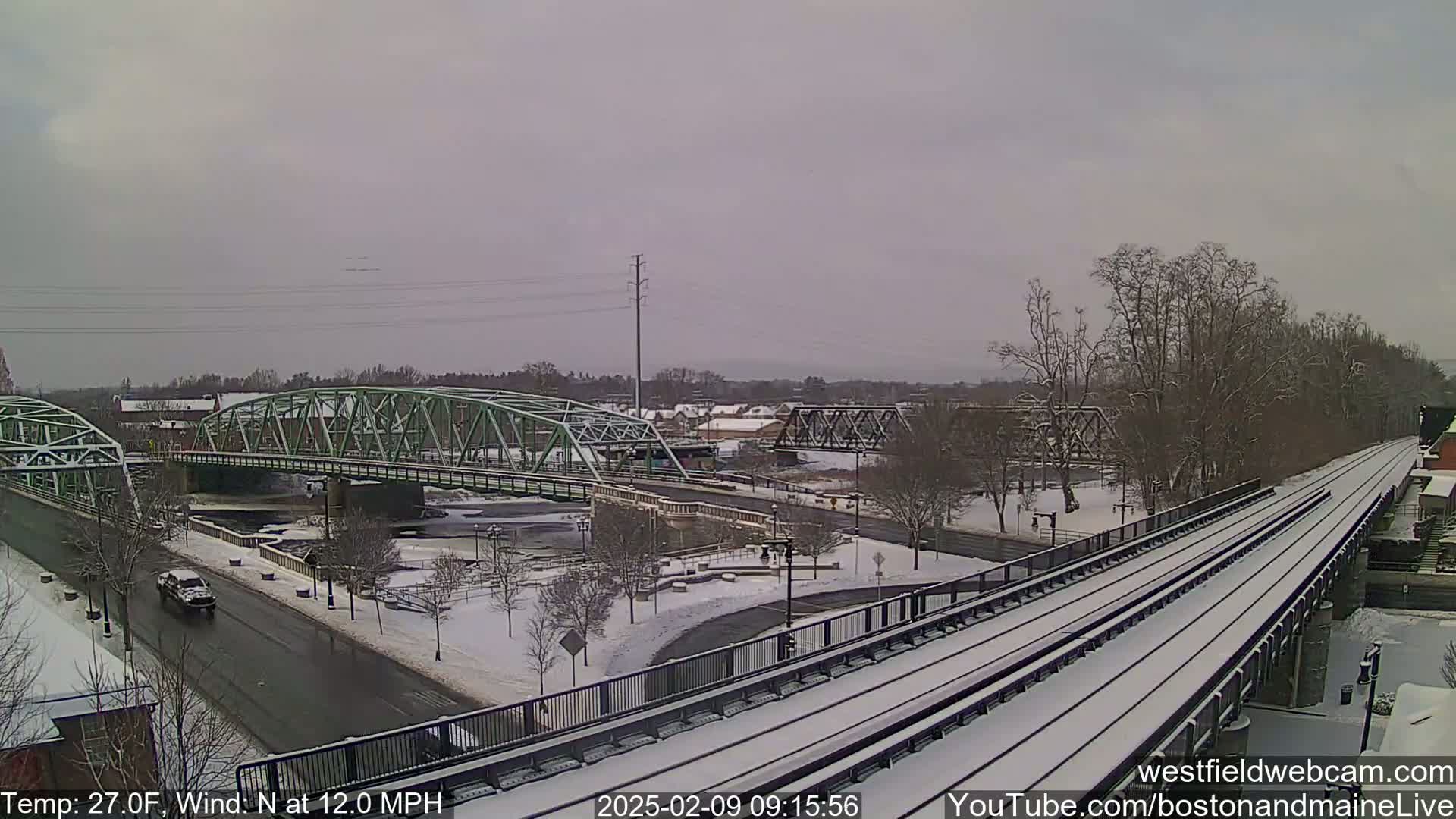 A snow-covered train track runs alongside a road and river, with two bridges in the background under a gray sky.