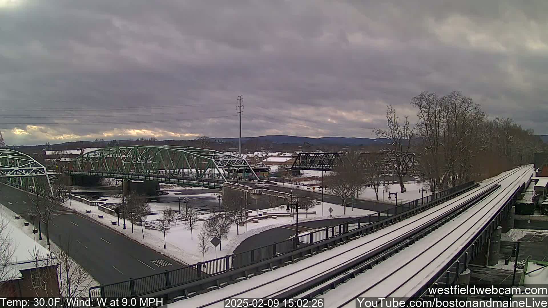 A snow-covered landscape features two bridges over a river, with a train track in the foreground under an overcast sky.