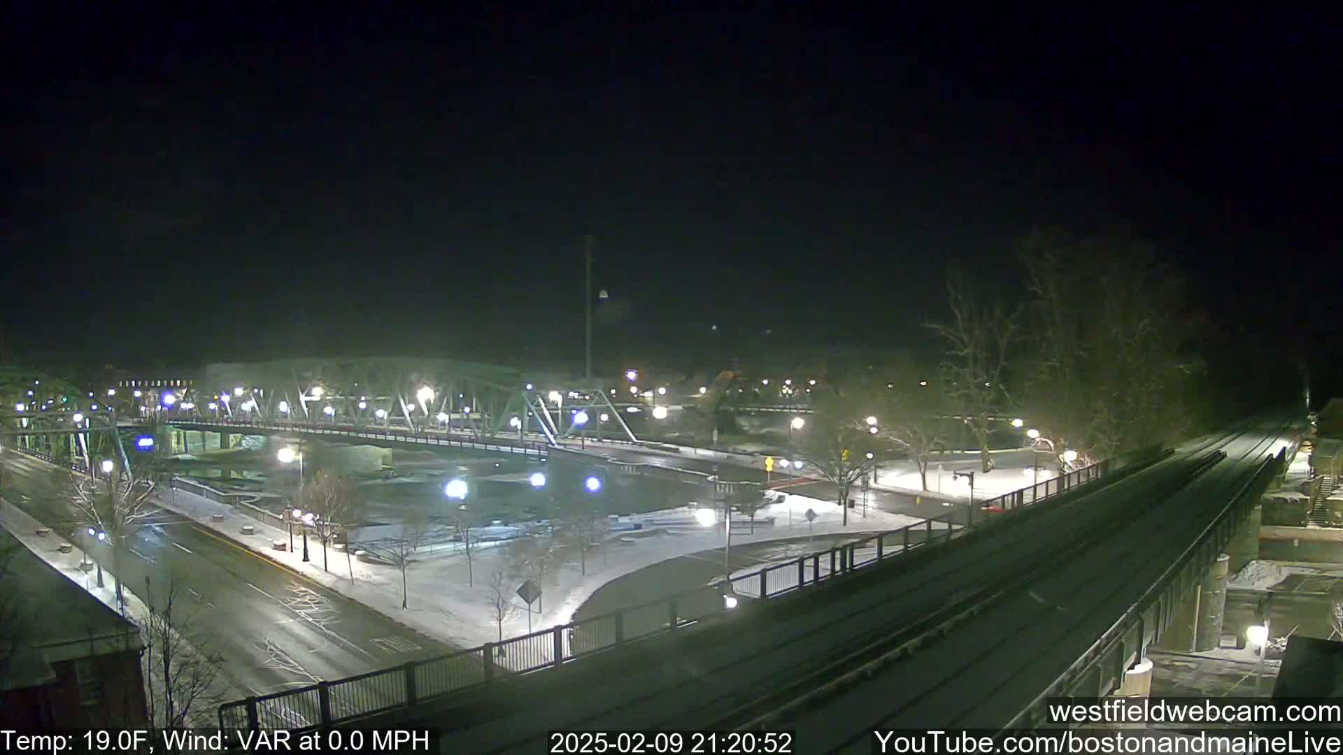 A nighttime view of a snow-covered bridge and roadway, with a calm wind and a temperature of 19 degrees Fahrenheit.