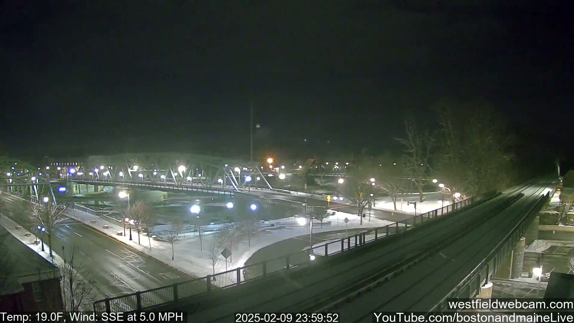 A snow-covered bridge and roadway at night, with a train track in the foreground under a dark sky.