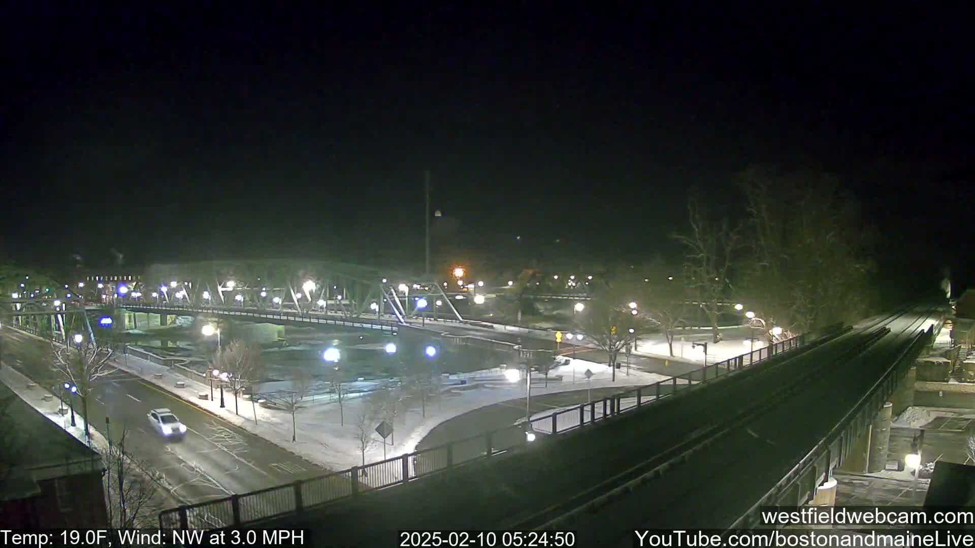 A snow-covered bridge and roadway are illuminated at night, with a light snowfall and a northwest wind.