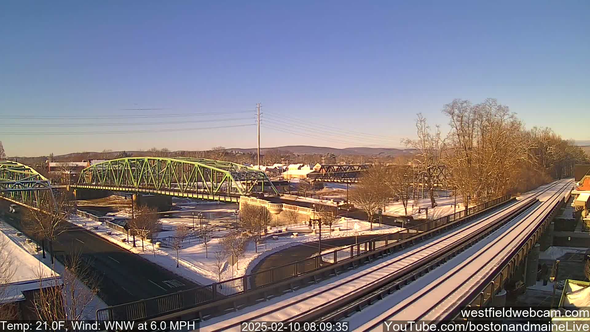 A snow-covered landscape with two bridges, one green and one dark brown, beside a train track on a sunny day.