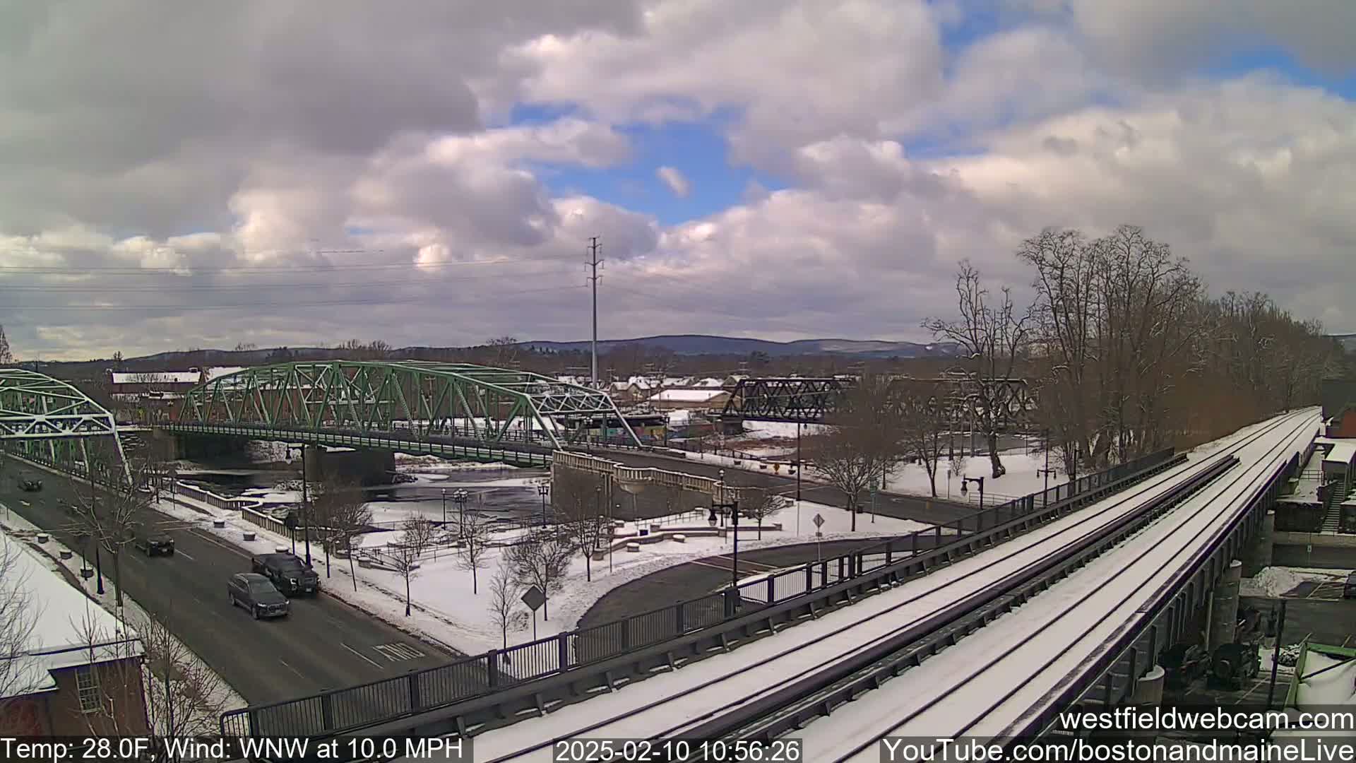 A snow-covered landscape with two bridges over a river, a train track, and several cars on a road, under a partly cloudy sky.