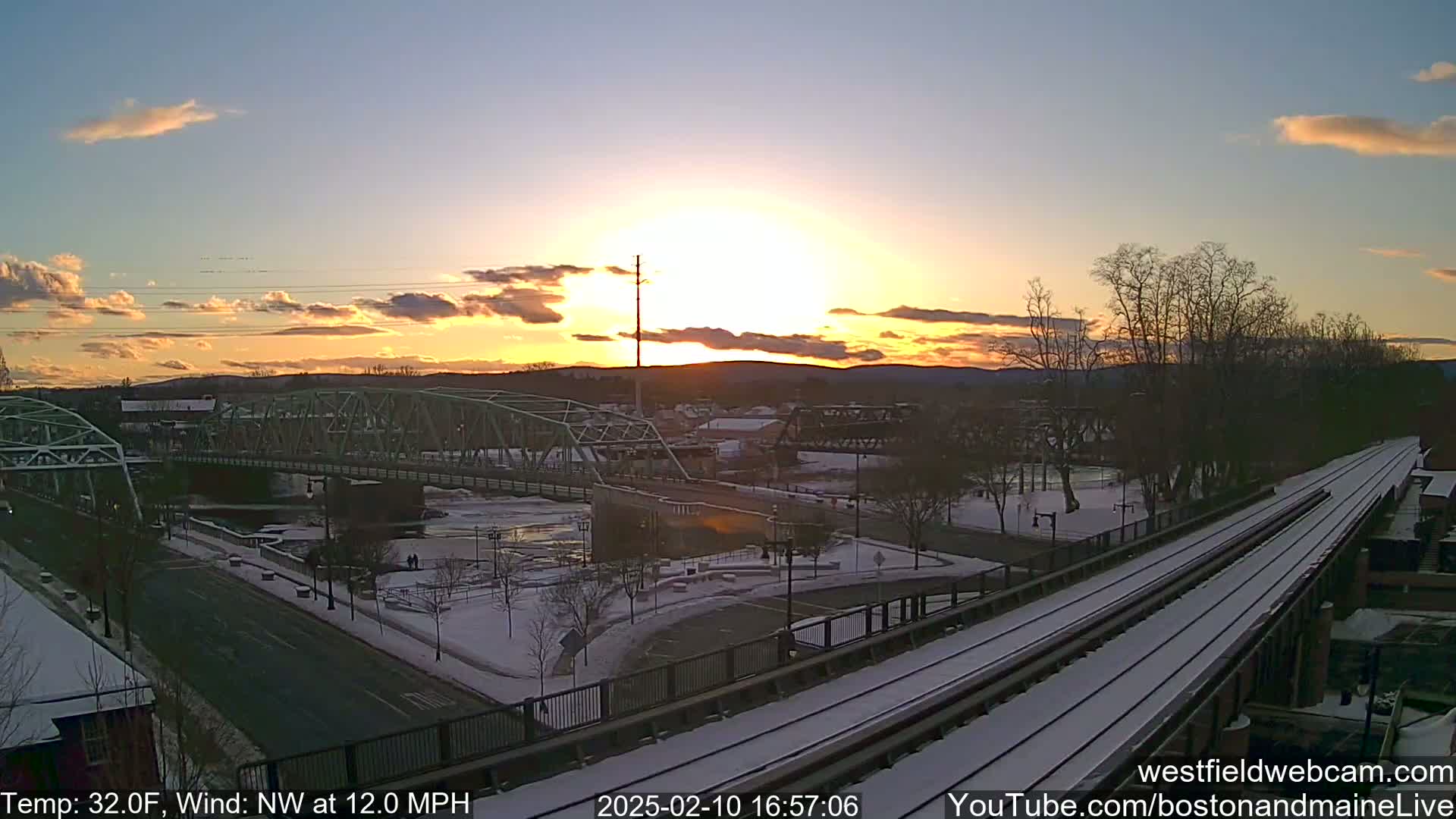 A snow-covered town scene at sunset, featuring a bridge over a river, with a train track in the foreground and a 32°F temperature with a 12 mph northwest wind.