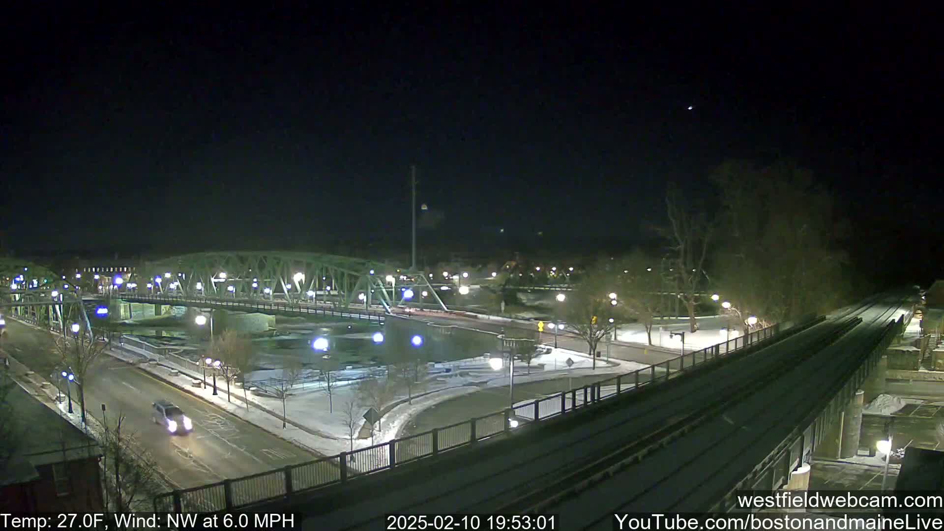 A nighttime view of a snow-covered bridge and roadway, with a train track and sparsely lit park visible, under a clear, dark sky.