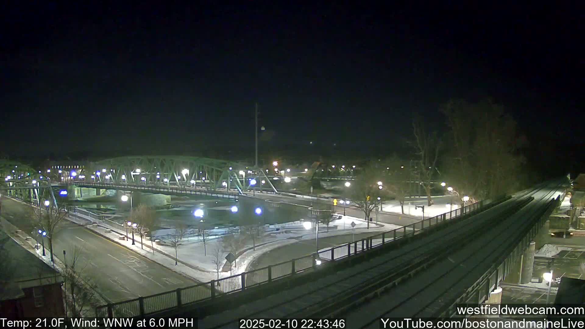 A nighttime view of a snow-covered bridge and road, with a train track alongside, under a clear, dark sky and a temperature of 21°F, with a wind from the west-northwest at 6.0 mph.