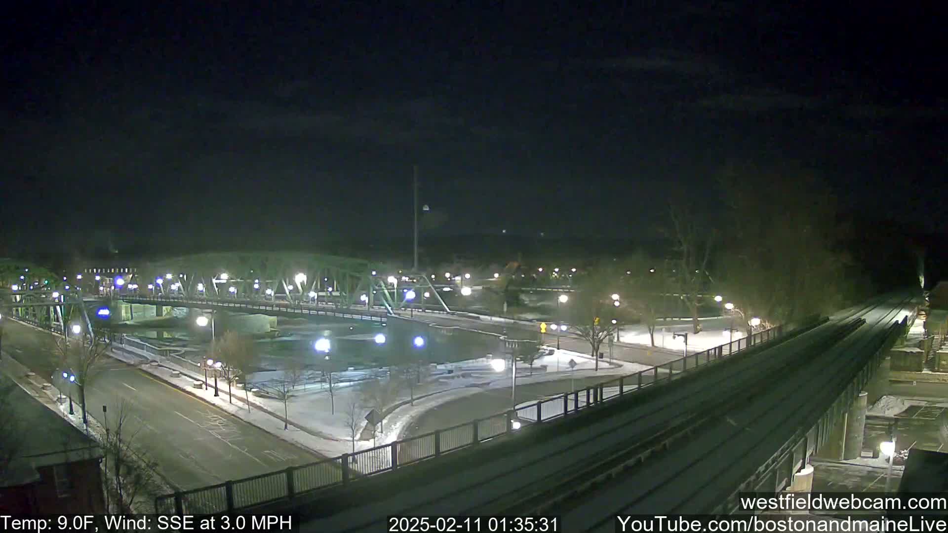 A nighttime view of a snow-covered bridge and roadway with a train track in the foreground, under a clear sky.