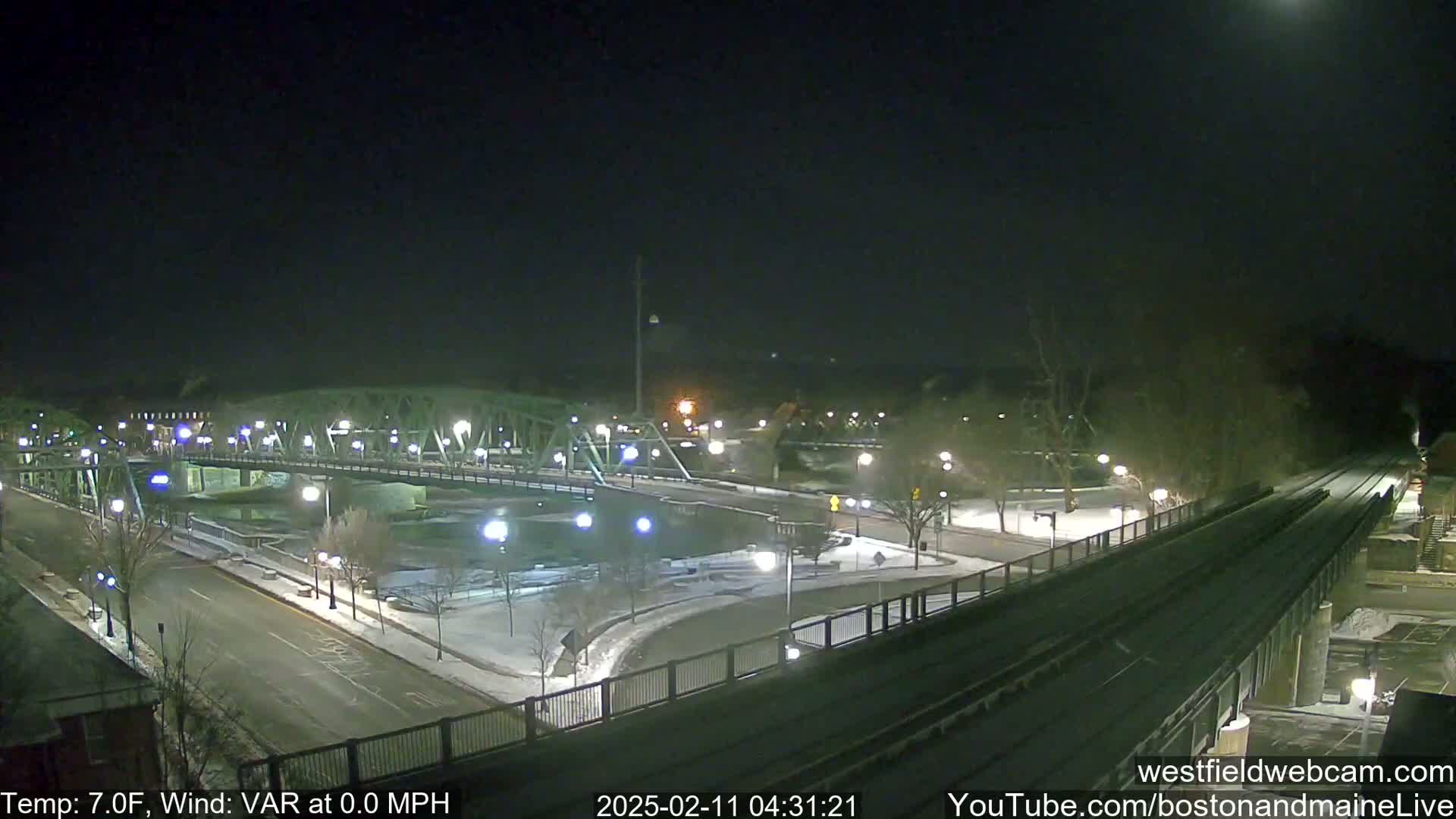 A nighttime view of a snow-covered bridge and roadway intersecting near a river, under a clear sky with a bright moon.