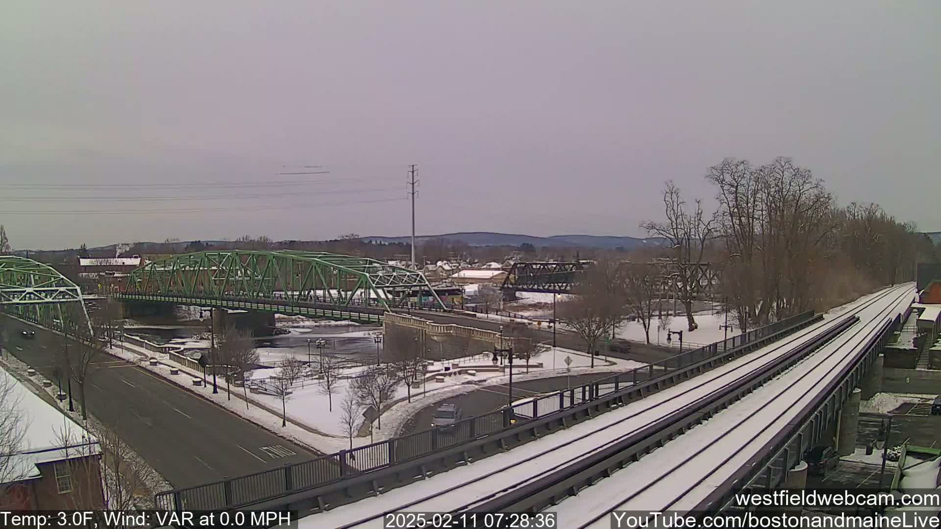 A snow-covered urban scene with a green bridge over a river, train tracks, and bare trees under a cloudy sky.