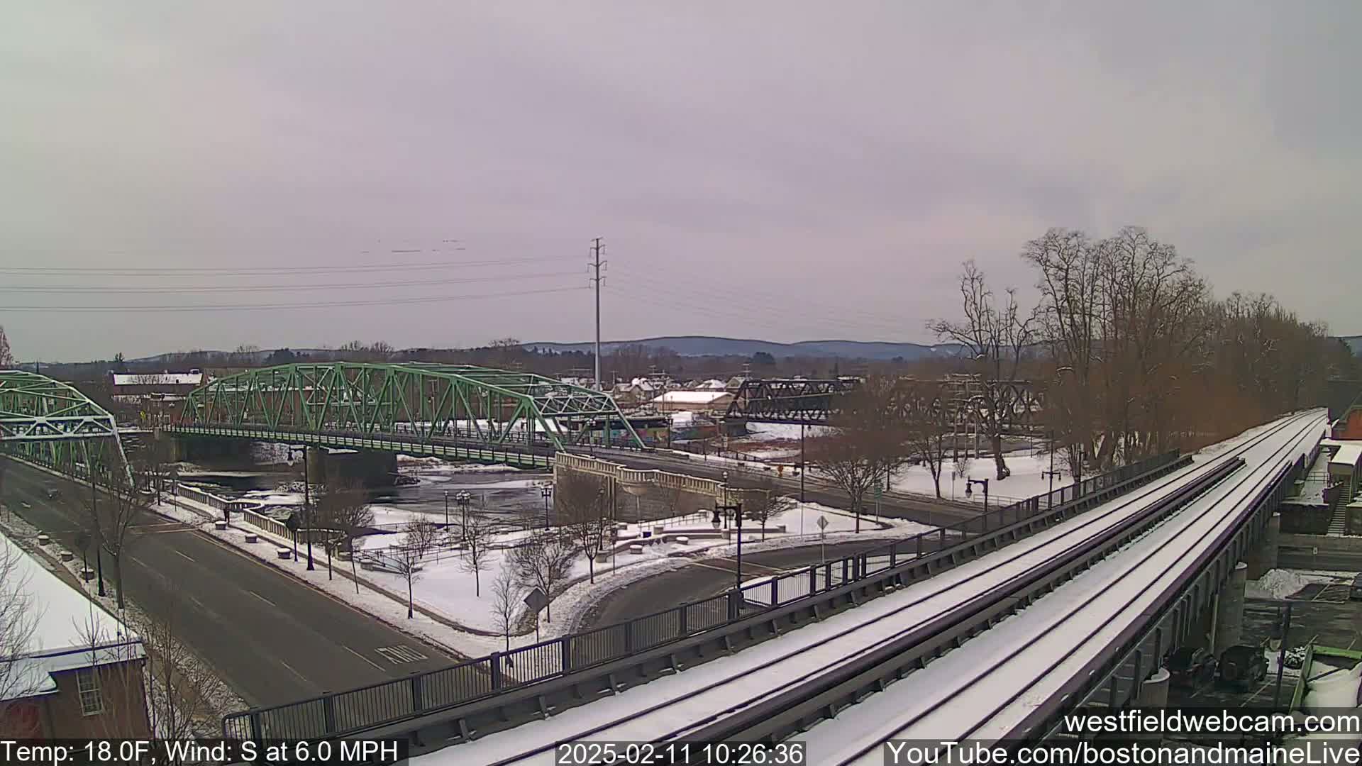 A snow-covered landscape shows two bridges over a river, with a train track running through a park-like area, under a cloudy sky with a south wind at 6 mph and a temperature of 18 degrees Fahrenheit.
