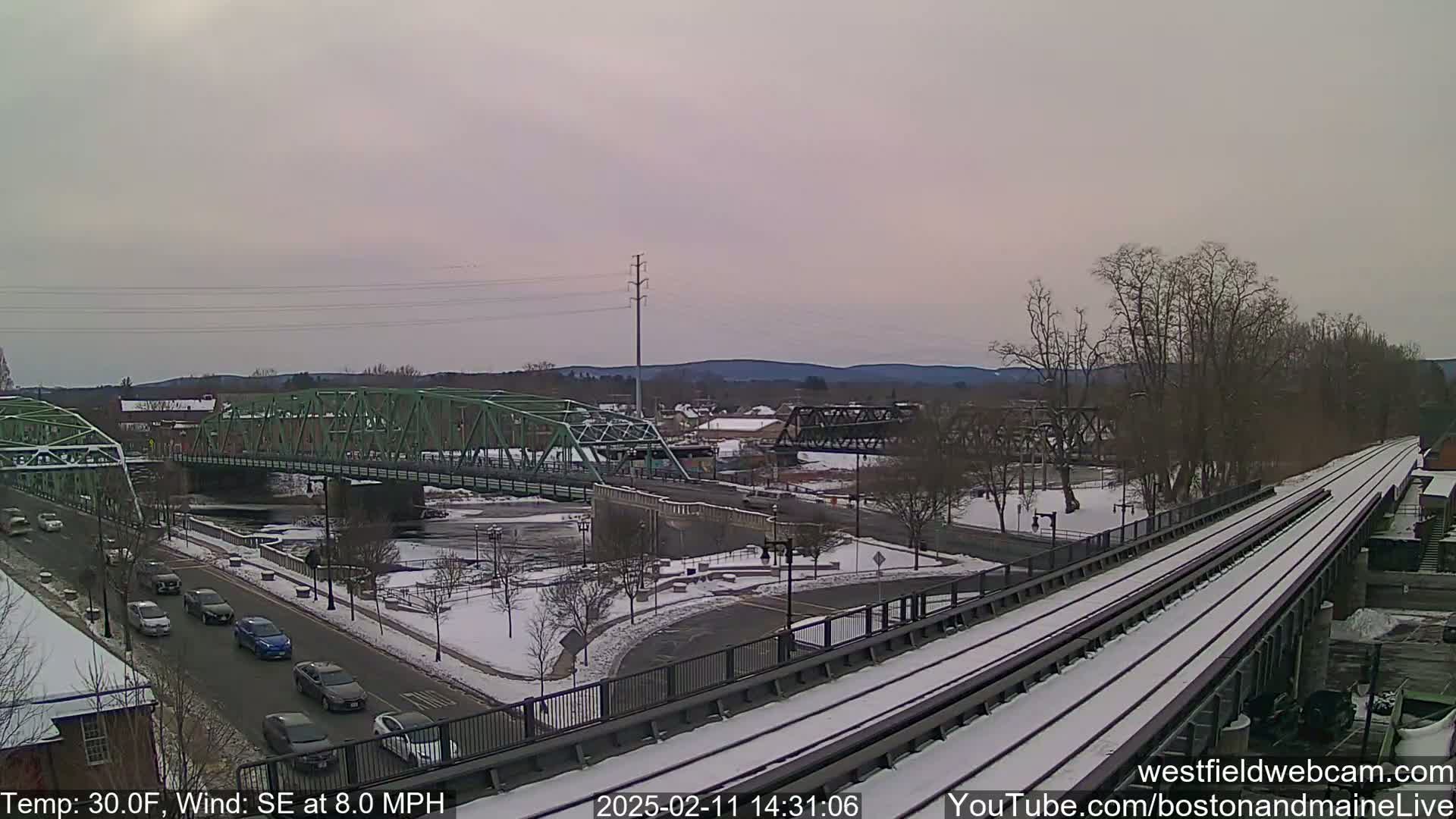 A snow-covered town scene with two bridges over a river, viewed from an elevated perspective showing train tracks in the foreground, under a cloudy sky.