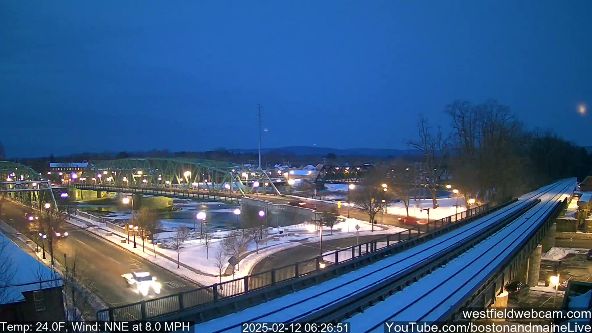 A snow-covered town at twilight, featuring a green bridge over a river, a roadway with sparse traffic, and elevated train tracks.
