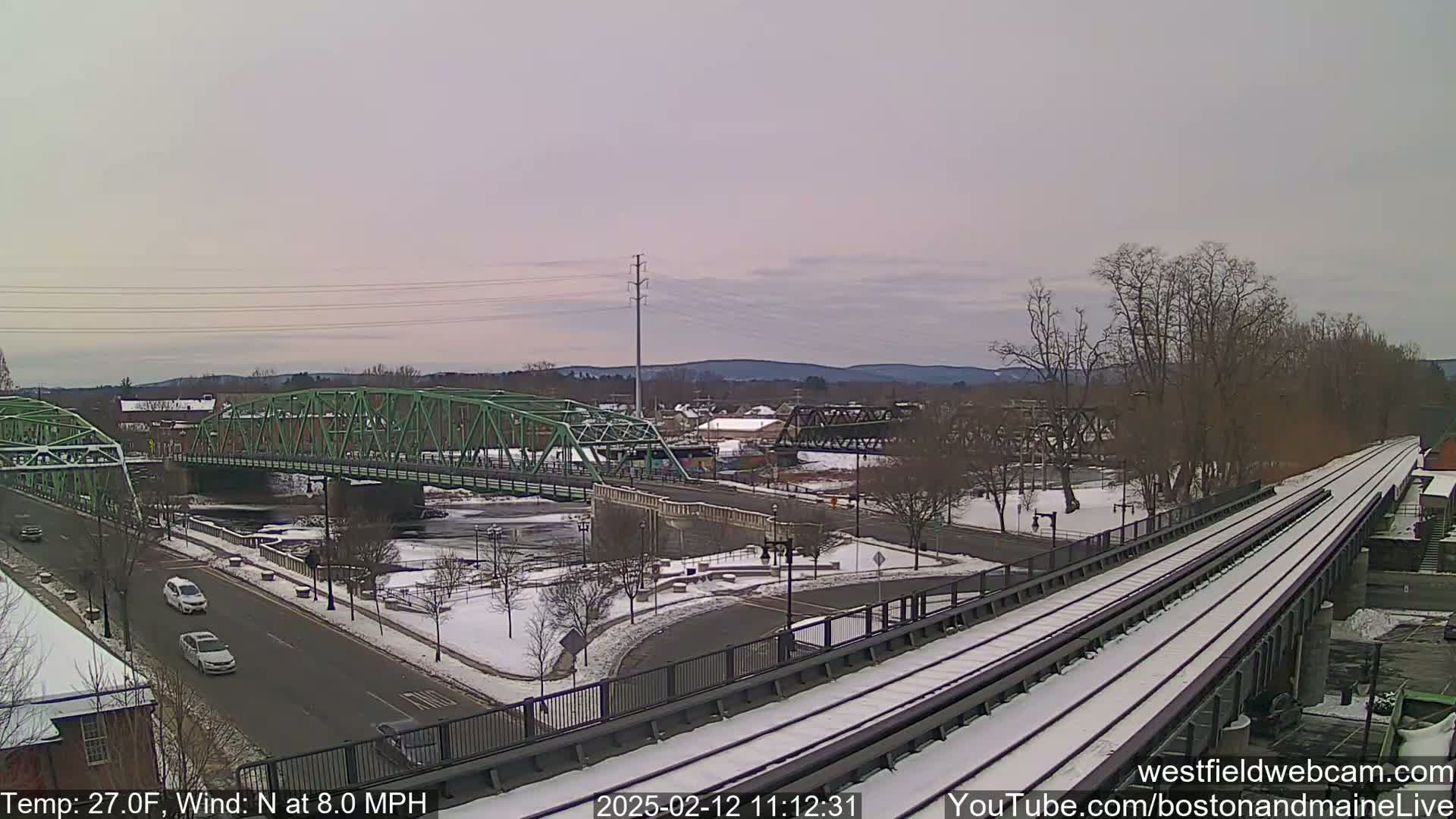 A snow-covered town scene with two bridges over a river, one green and one dark metal, alongside a train track on an overcast day.