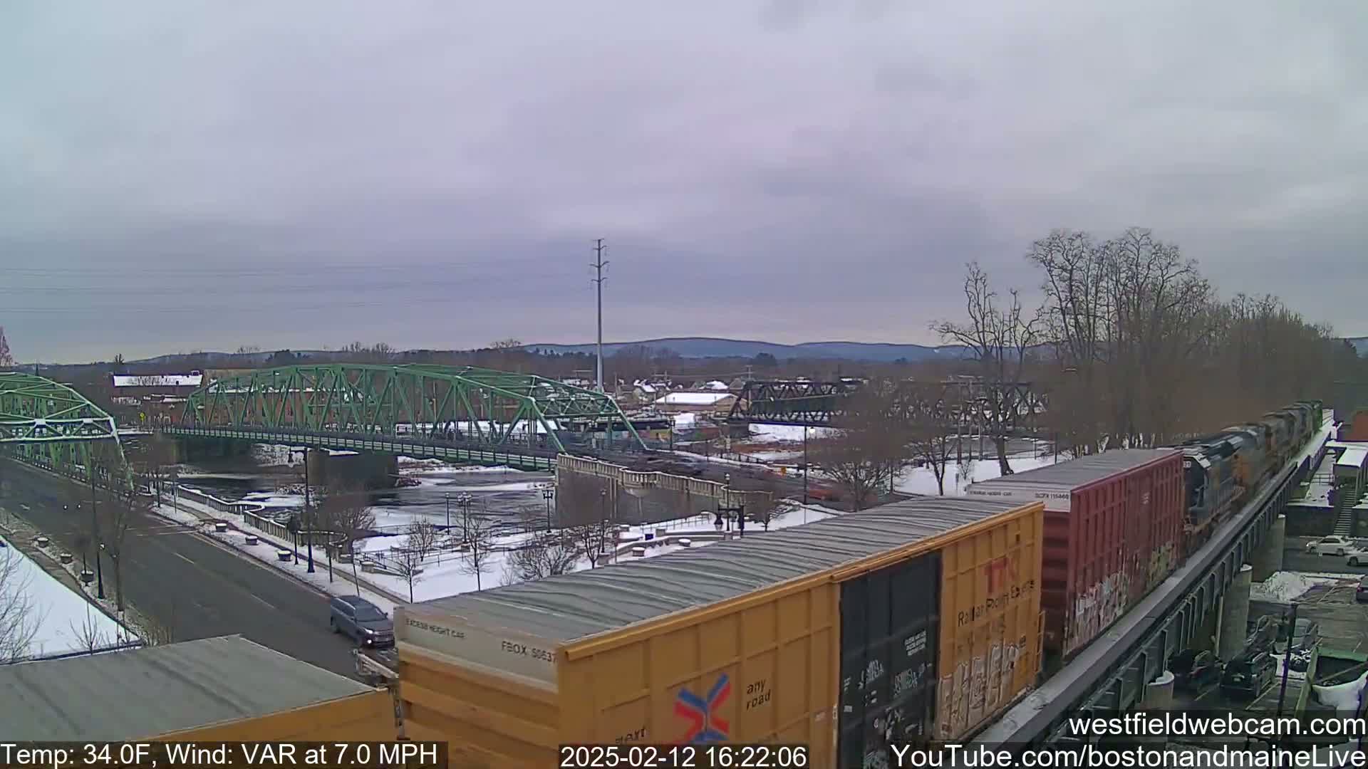 A freight train passes over a bridge next to a river, with a partly cloudy sky and light snow cover on the ground.